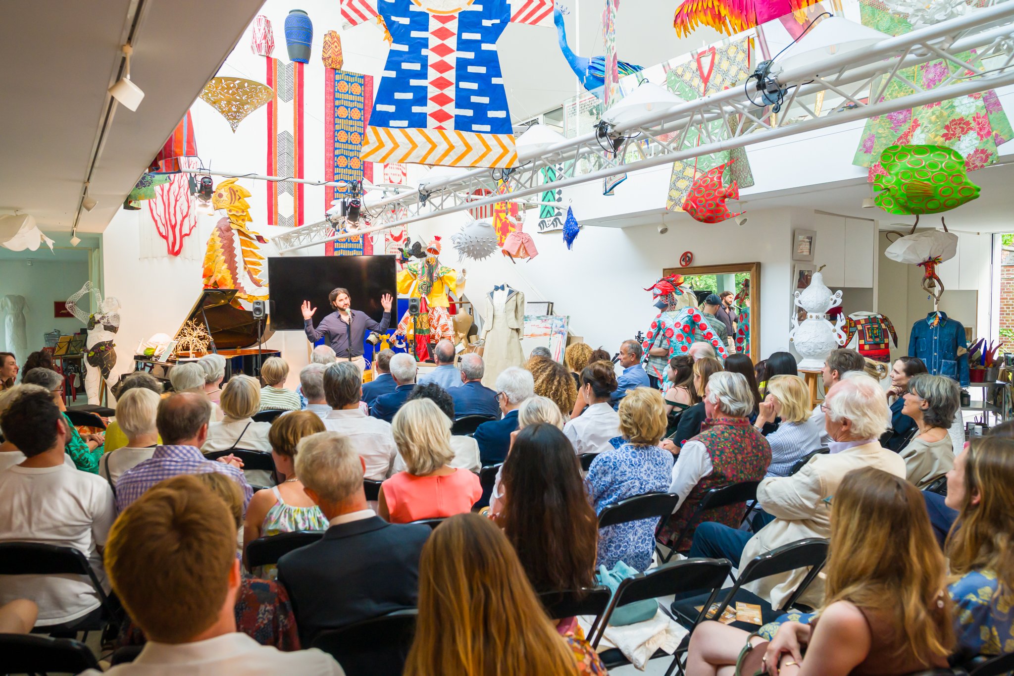 Une audience assise lors d'un événement culturel ou artistique, avec des décorations colorées suspendues au plafond, dont des parapluies, des lézards en papier et d'autres objets artistiques, dans une pièce bien éclairée avec un présentateur parlant 