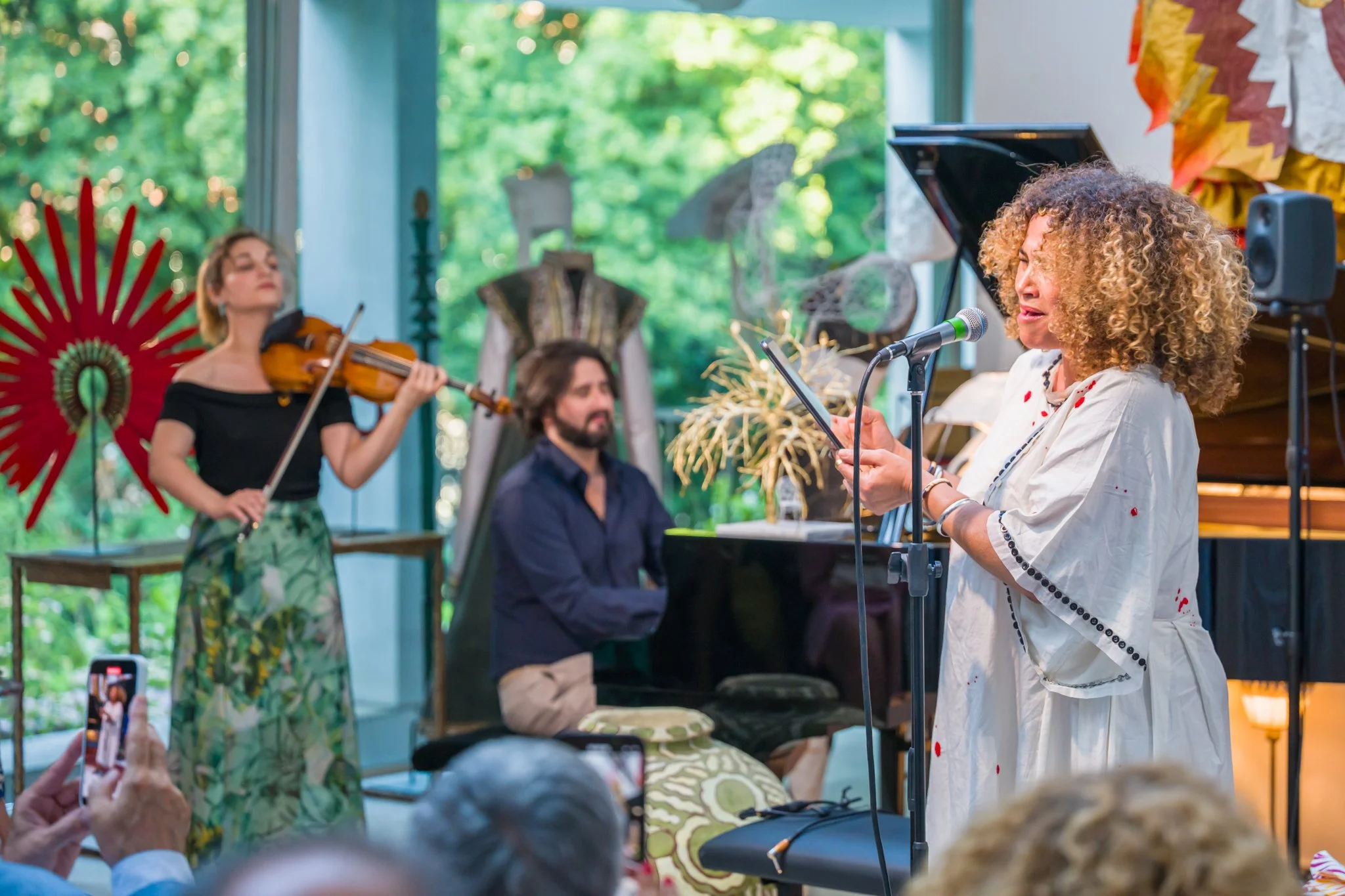 Une femme chante en lisant un texte, avec un trio de musiciens en arrière-plan : une violoniste, un pianiste, et des décorations colorées, lors d'une performance en intérieur près d'une fenêtre donnant sur un espace vert.