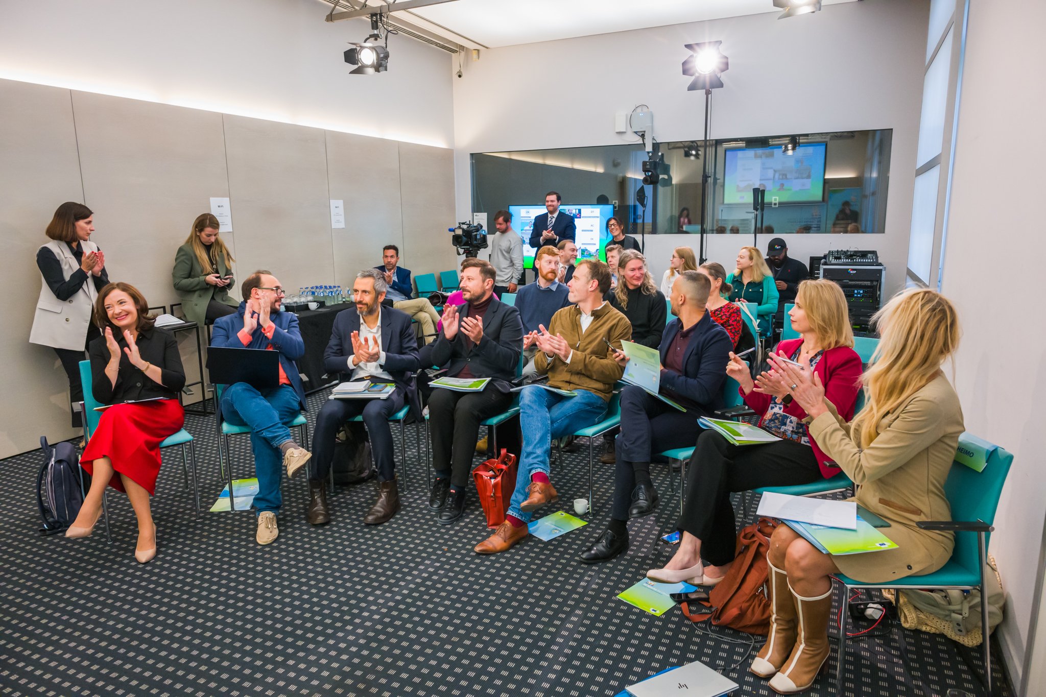 Groupe de personnes applaudissant lors d'un événement en salle, avec des chaises et du matériel audiovisuel.