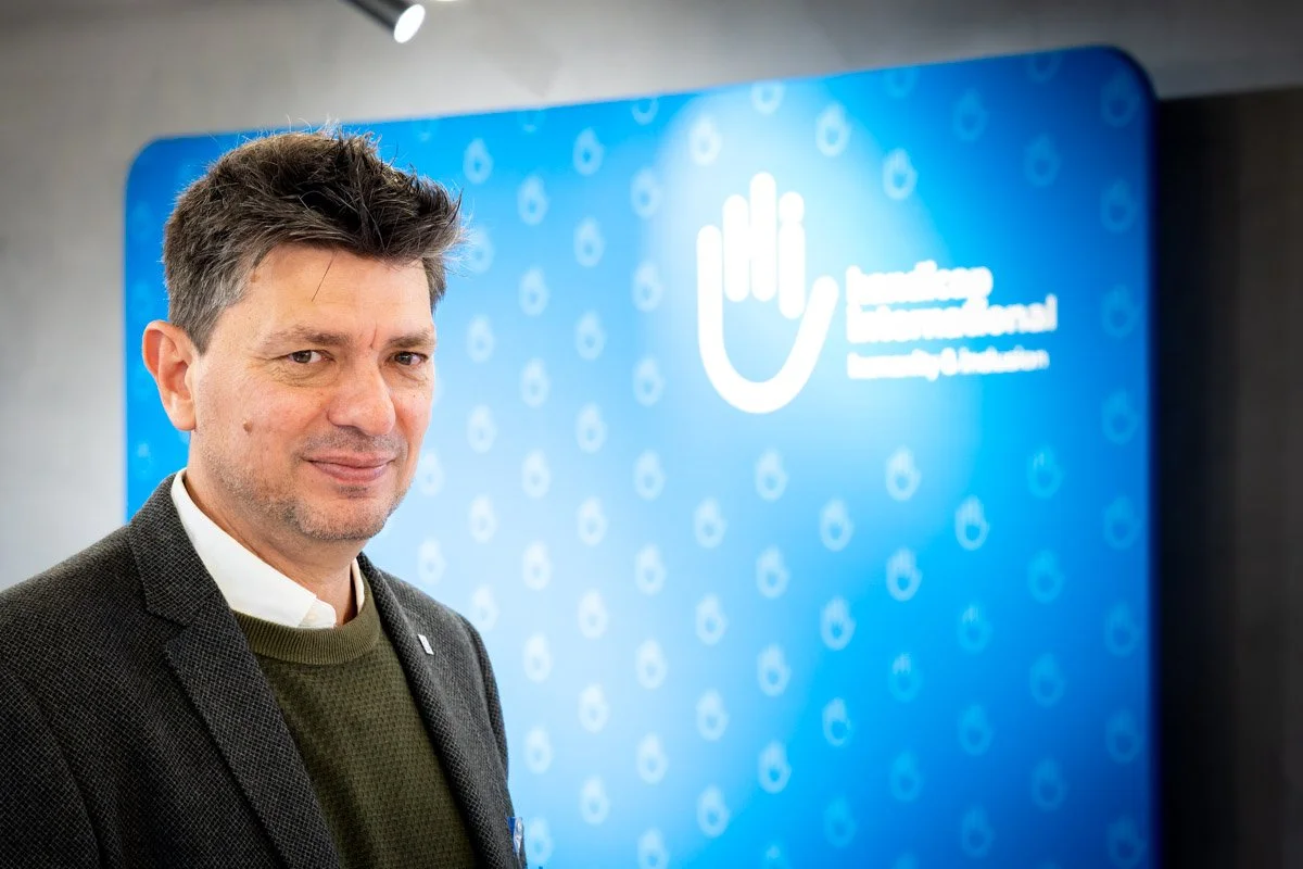 Un homme souriant en costume, se tenant devant un panneau bleu avec un logo et des textes en français suisent de la main.