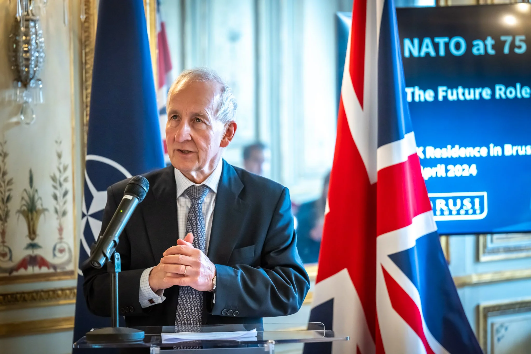 A costumed speaker speaks at a microphone, with NATO and UK flags in the background, at an event commemorating NATO's 75th anniversary.