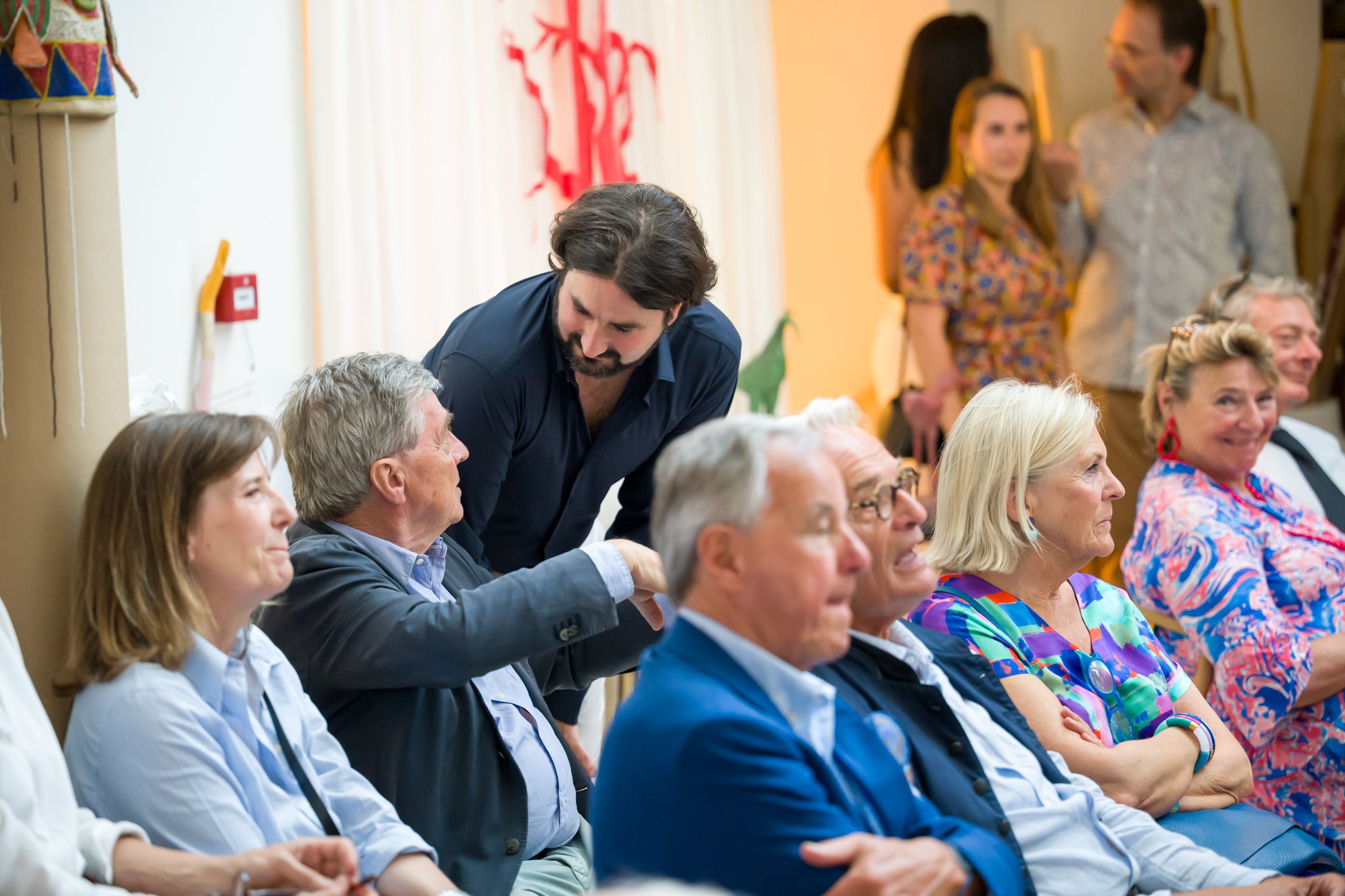 Groupe de personnes âgées assises dans une salle, conversation animée avec une personne debout, fond chaleureux avec décoration colorée.