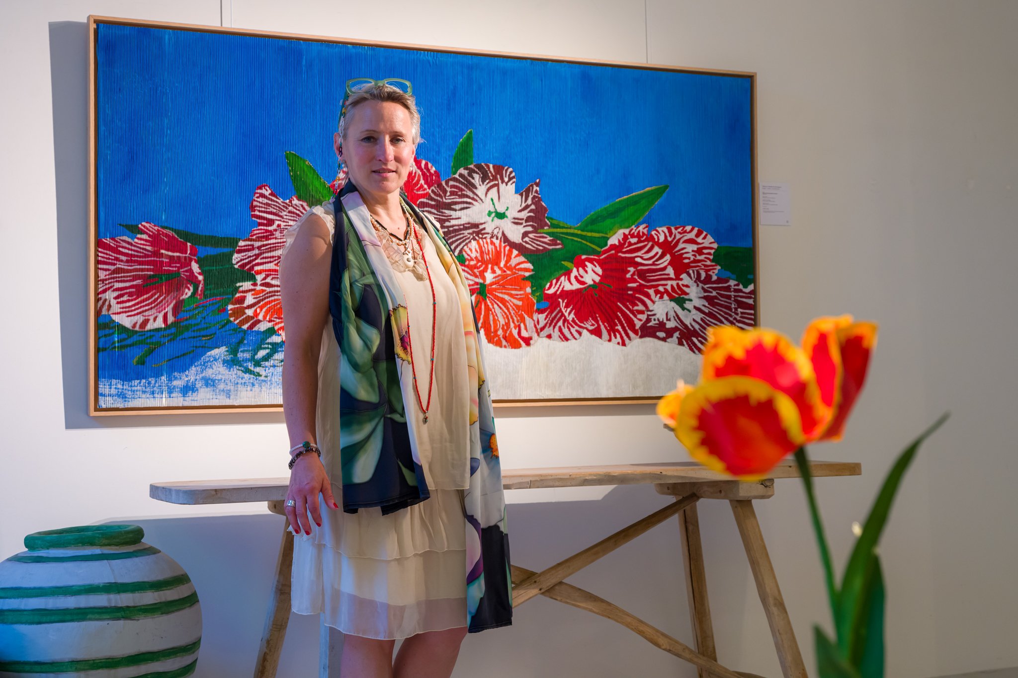Femme posant devant une œuvre d'art florale dans une galerie, avec un vase coloré en premier plan.