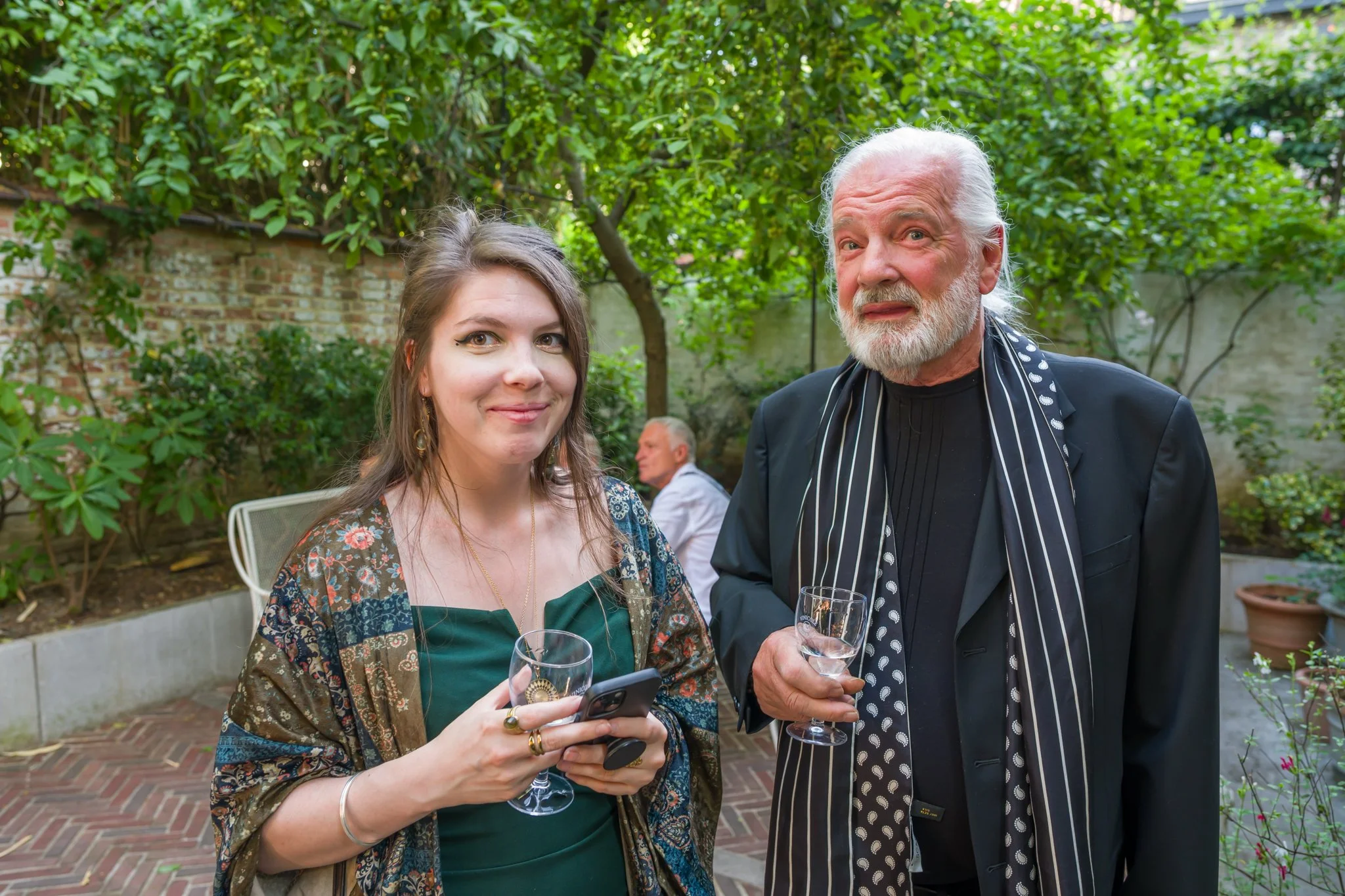Une jeune femme et un homme âgé portant une tenue élégante, tenant des verres à vin lors d'une fête en extérieur, avec un jardin en arrière-plan.