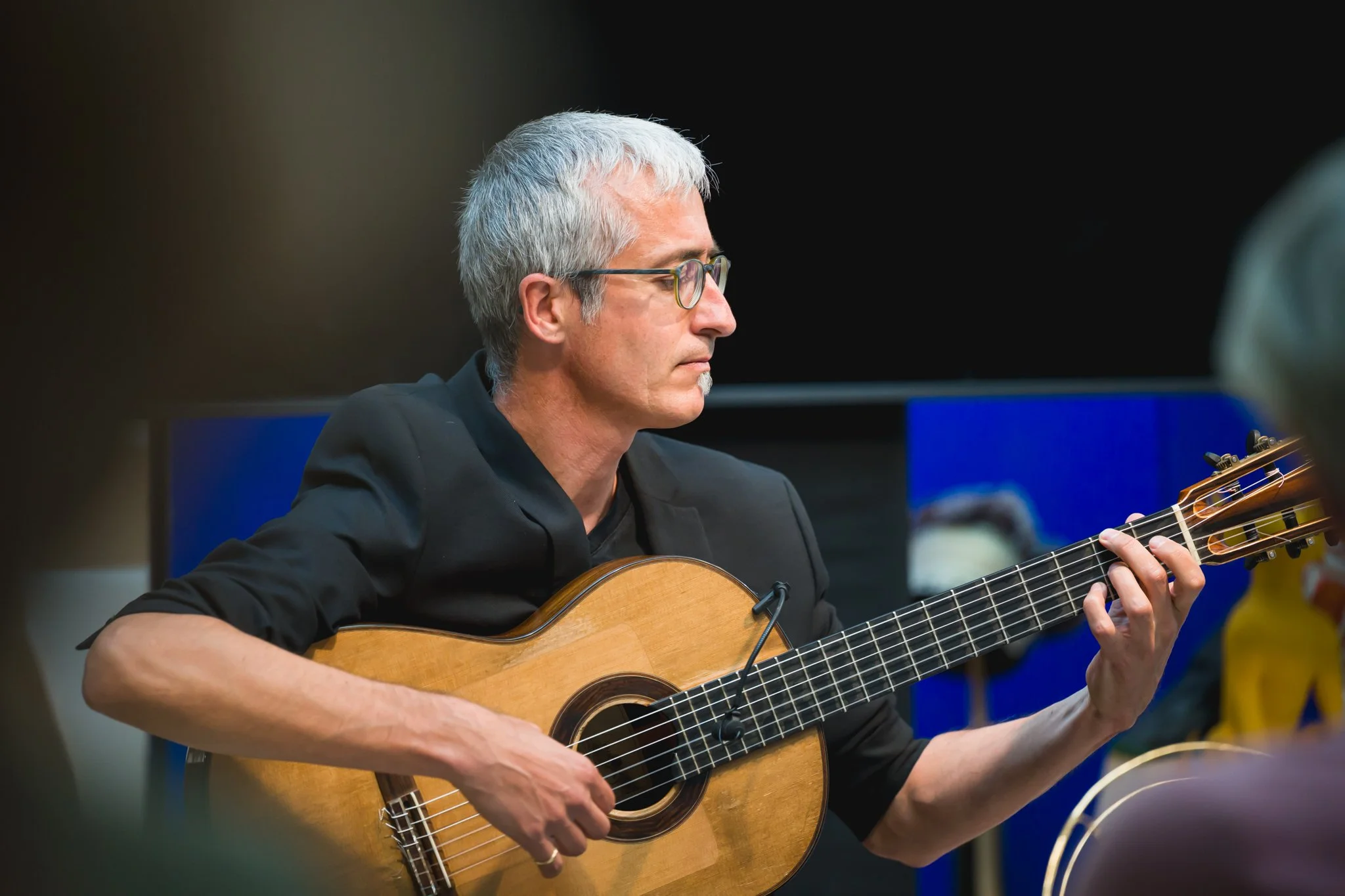 Un homme aux cheveux gris, portant des lunettes et une veste noire, joue de la guitare acoustique lors d'une performance ou d'un rassemblement.