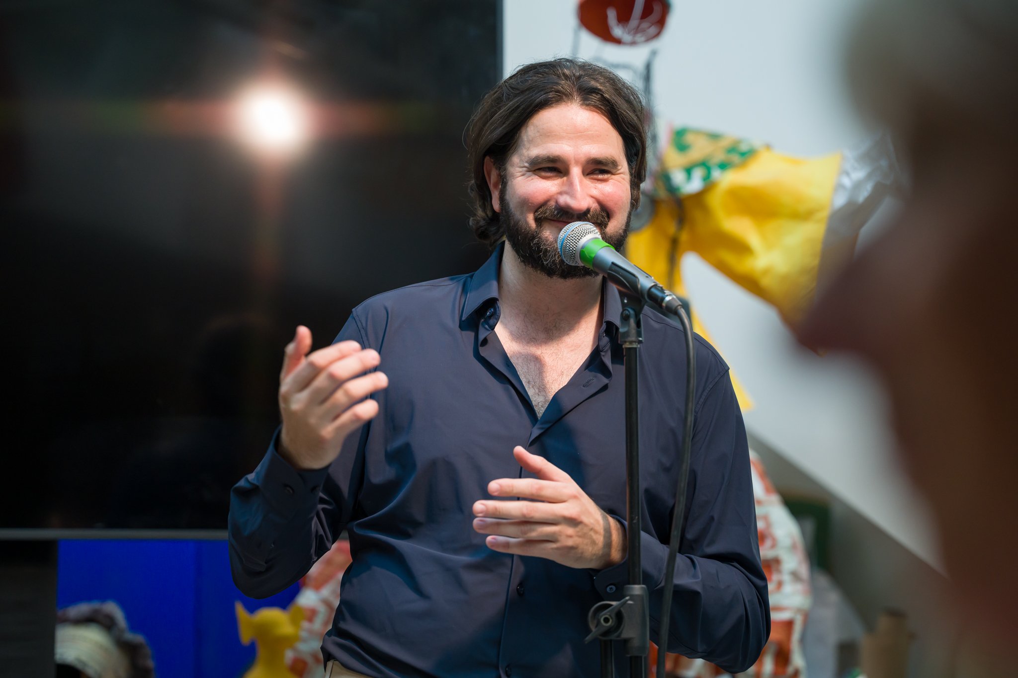 Un homme avec des cheveux bruns, barbe, portant une chemise bleue, parle dans un microphone lors d'une présentation ou d'un événement, souriant, avec un fond contenant un grand écran noir et un bouquet de fleurs.