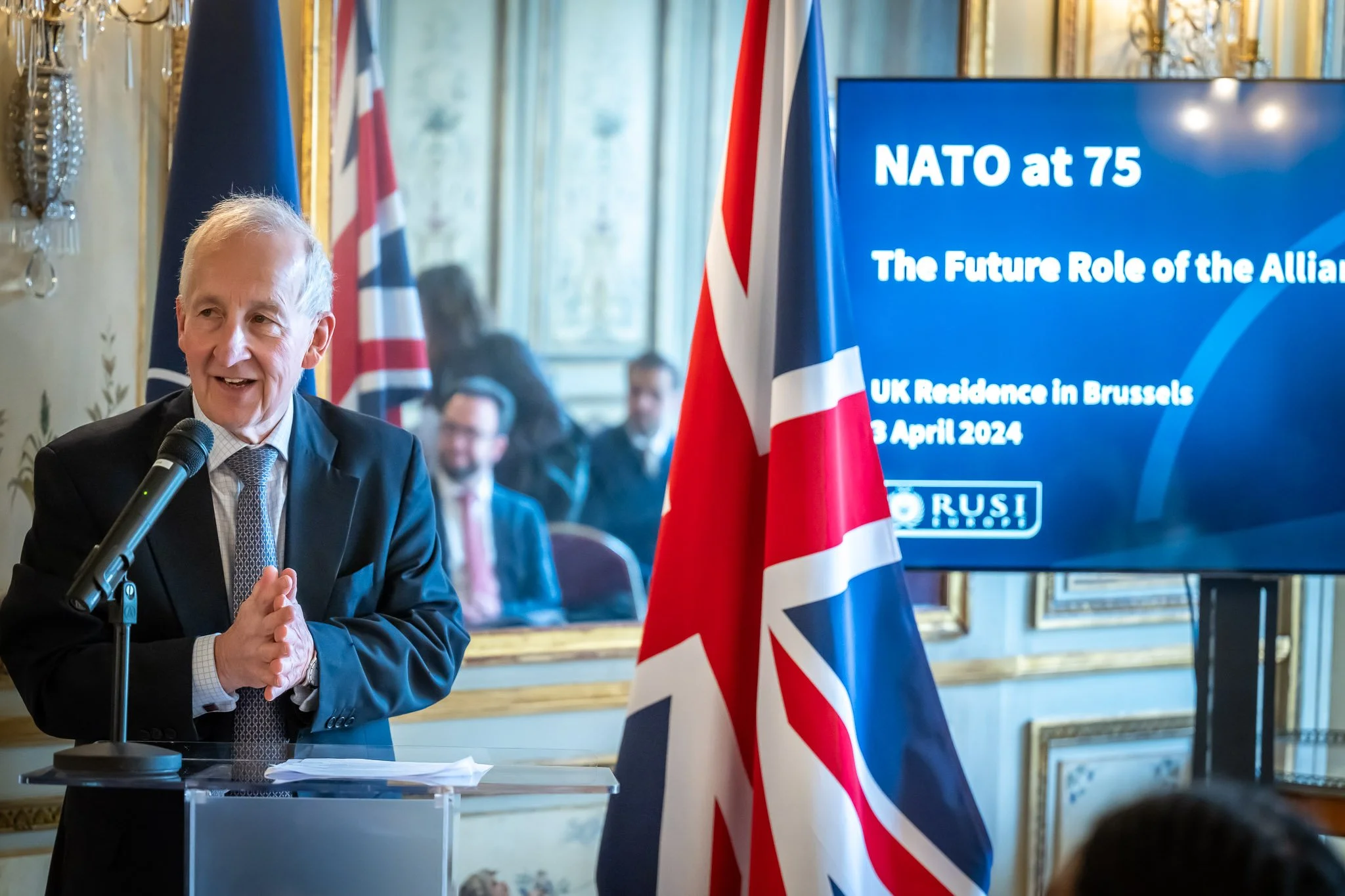 Politician speaking at a podium, British flag and screen displaying "NATO at 75, The Future Role of the Alliance, UK Residence in Brussels, 8 April 2024".