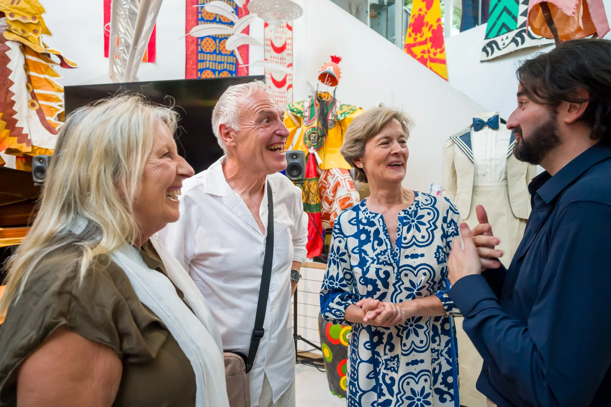 Groupe de quatre adultes souriants conversant dans un atelier d'art ou d'artisanat, avec des œuvres colorées en arrière-plan.