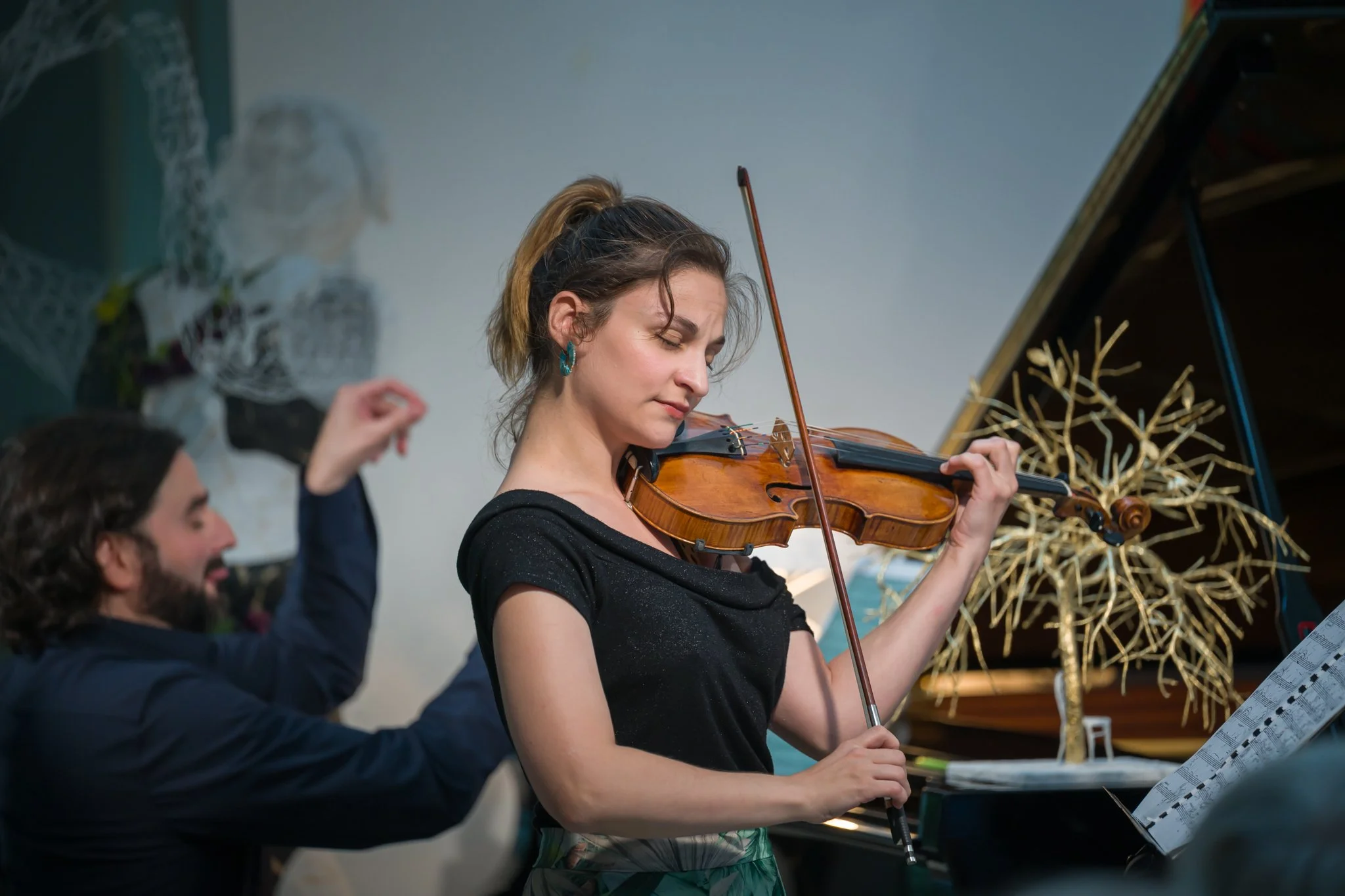 Une femme joue du violon en regardant une partition sur un piano, accompagnée d'un homme danser ou exécuter un mouvement de bras.