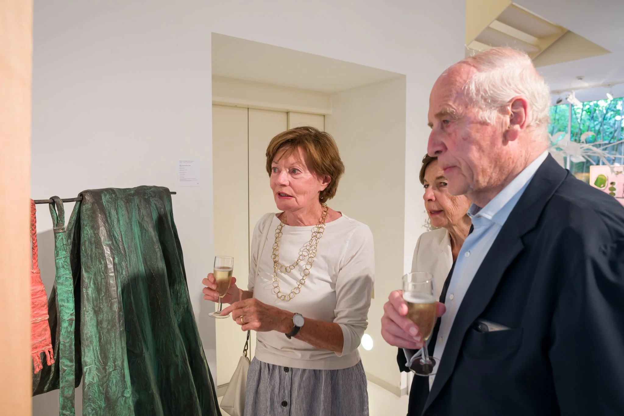 Trois personnes regardant des vêtements d'art en exposition lors d'un vernissage, dégustant du champagne.