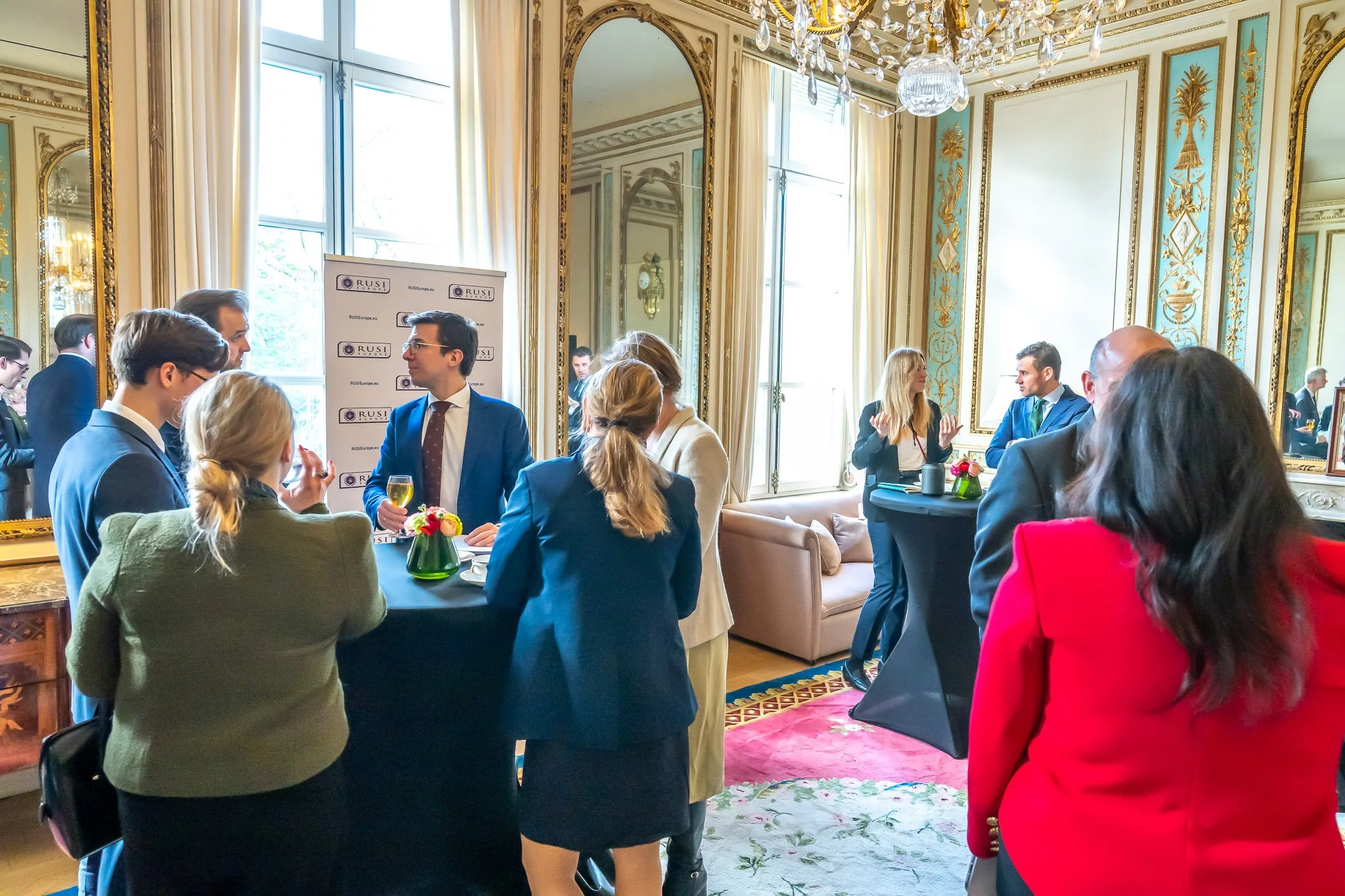 A group of formally dressed people chat in an elegant lounge with mirrors, a chandelier and curtains. Drinks and floral arrangements are laid out on small, high tables."}