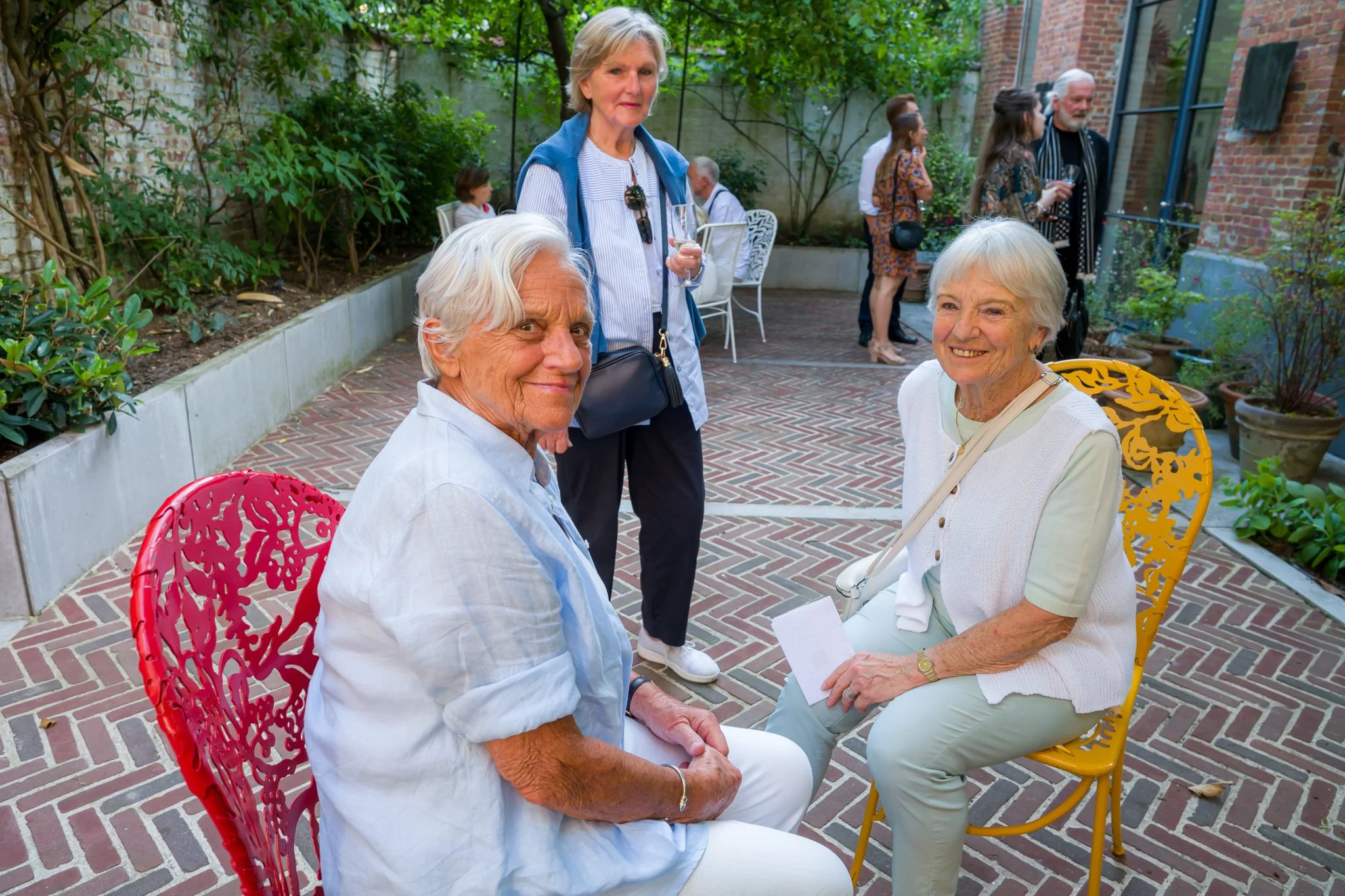 Trois femmes âgées profitant d'une rencontre en extérieur, deux assises face à face, la troisième debout derrière elles, dans un jardin avec plantes et mobilier, lors d'une occasion sociale en journée.