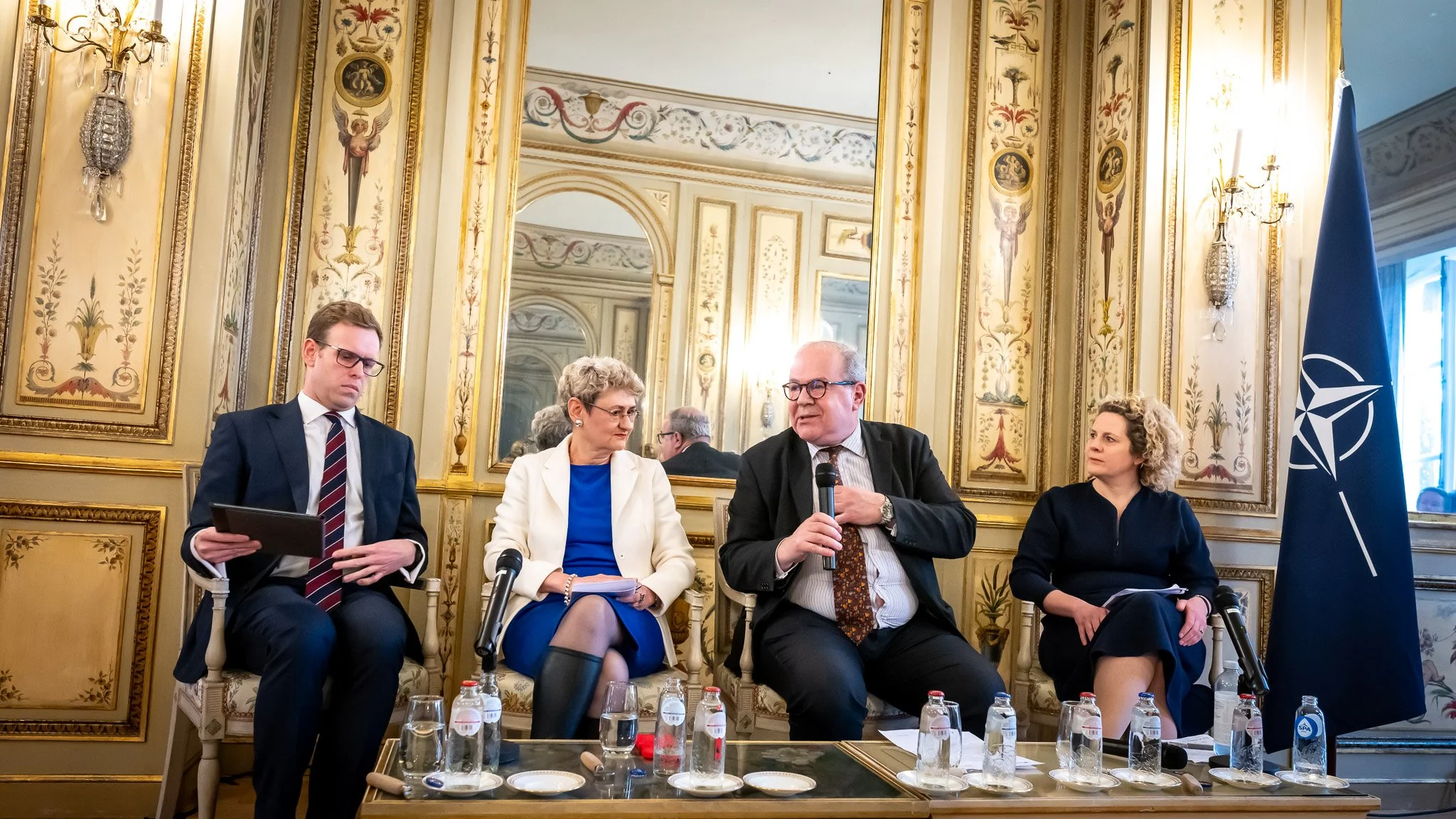 Four people sitting in an ornate room in front of a NATO flag, talking at a conference, with microphones and bottles of water on a table.