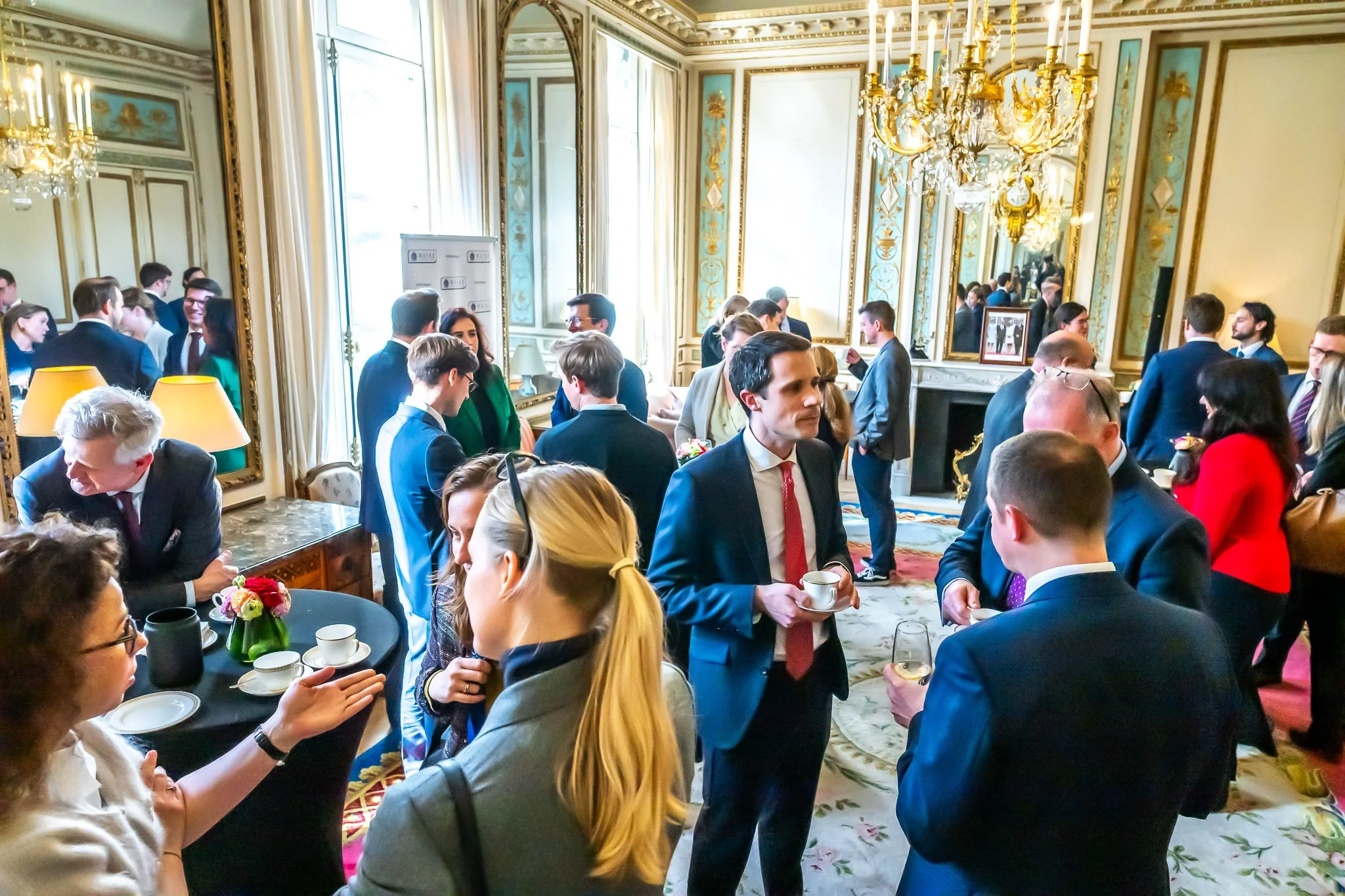 Formally dressed people chat at a social or business event in an elegant room with chandeliers and period mirrors.