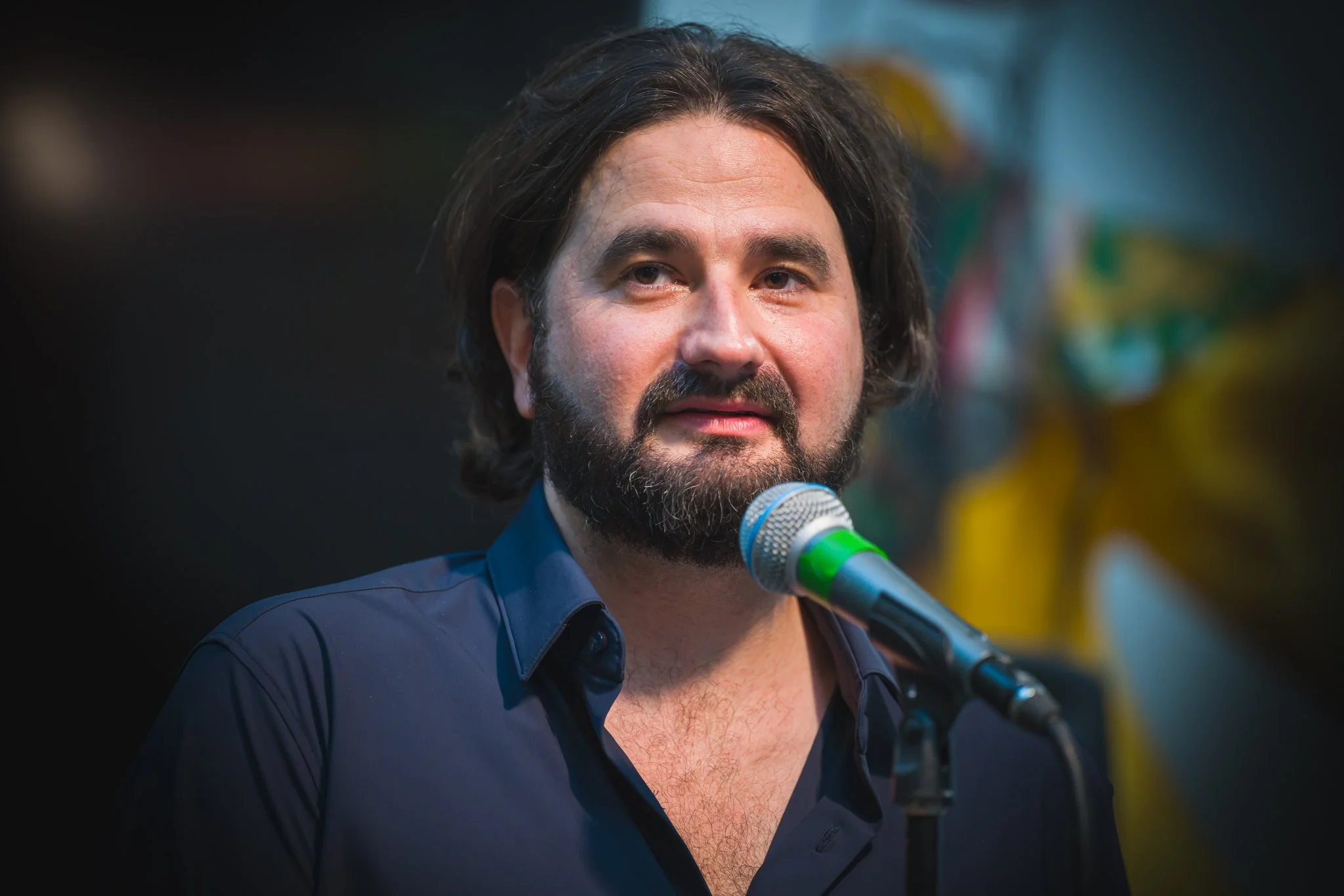 Un homme barbu, avec des cheveux mi-longs, portant une chemise bleue, parle devant un micro lors d'un discours ou d'une interview.