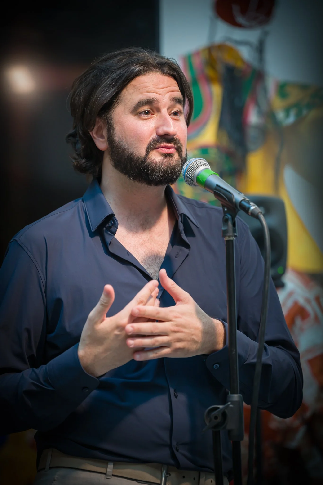 Homme avec une barbe et des cheveux longs, portant une chemise bleue foncée, parle devant un microphone lors d'une présentation ou d'une performance, avec un fond coloré et artistique.