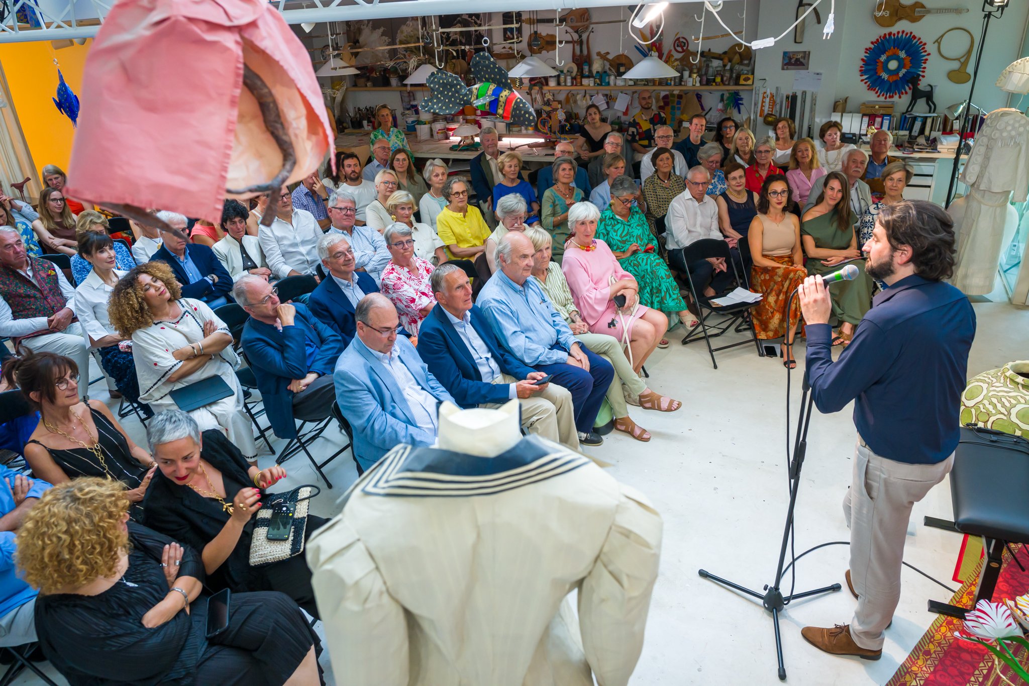 Un homme parle devant un groupe de personnes assises dans une salle, lors d'un événement ou d'une présentation. La salle est décorée avec des objets artistiques et des œuvres d'art.