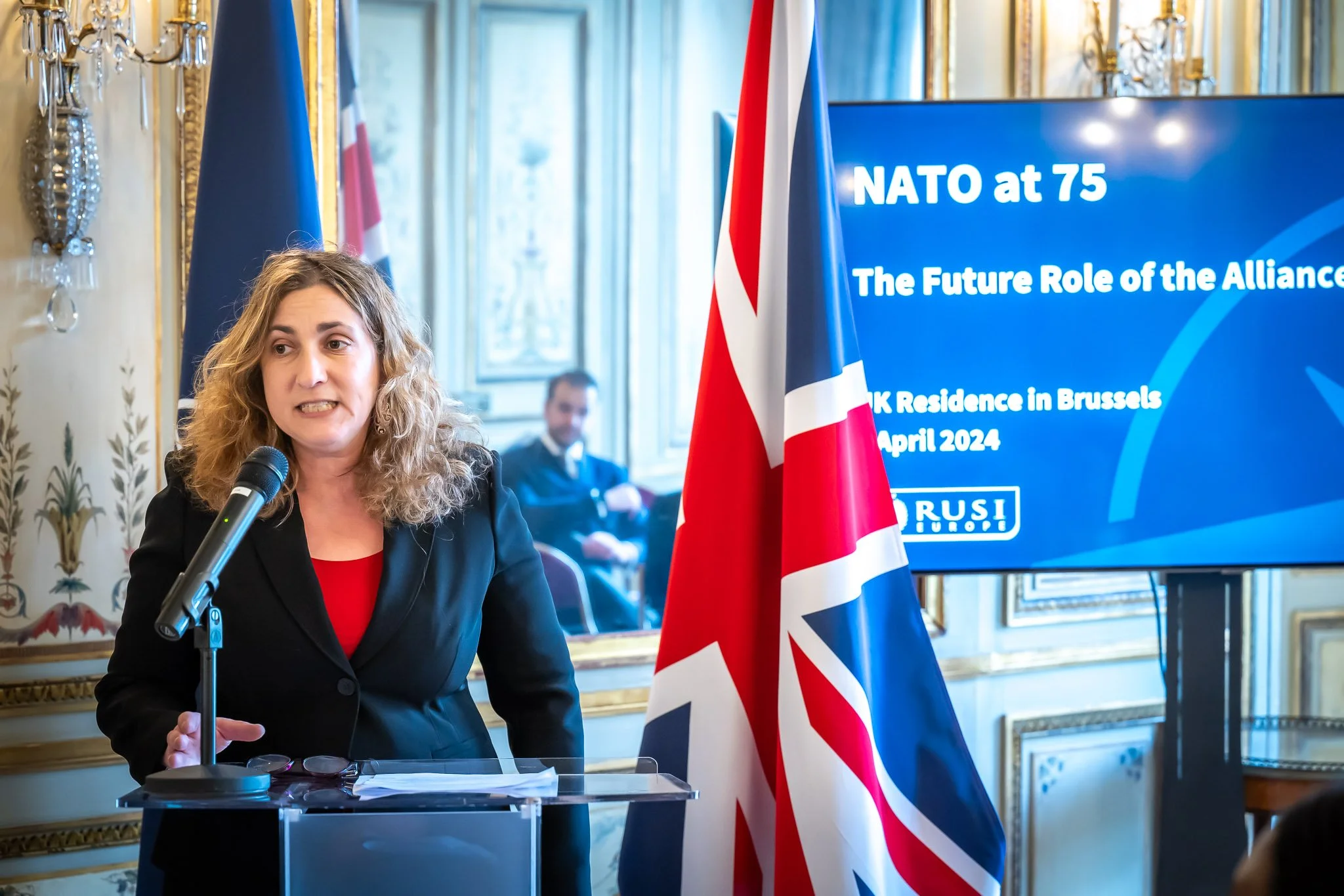 Woman speaking at microphone in ornate room, British flag in foreground, screen displaying "NATO at 75 - The Future Role of the Alliance", Union Jack visible, candelabra in background.