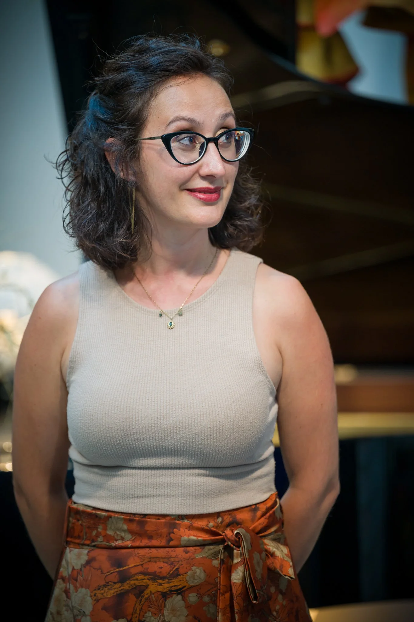 Une femme avec des lunettes, portant un débardeur beige et une jupe en tissu à motifs floraux, devant un piano.