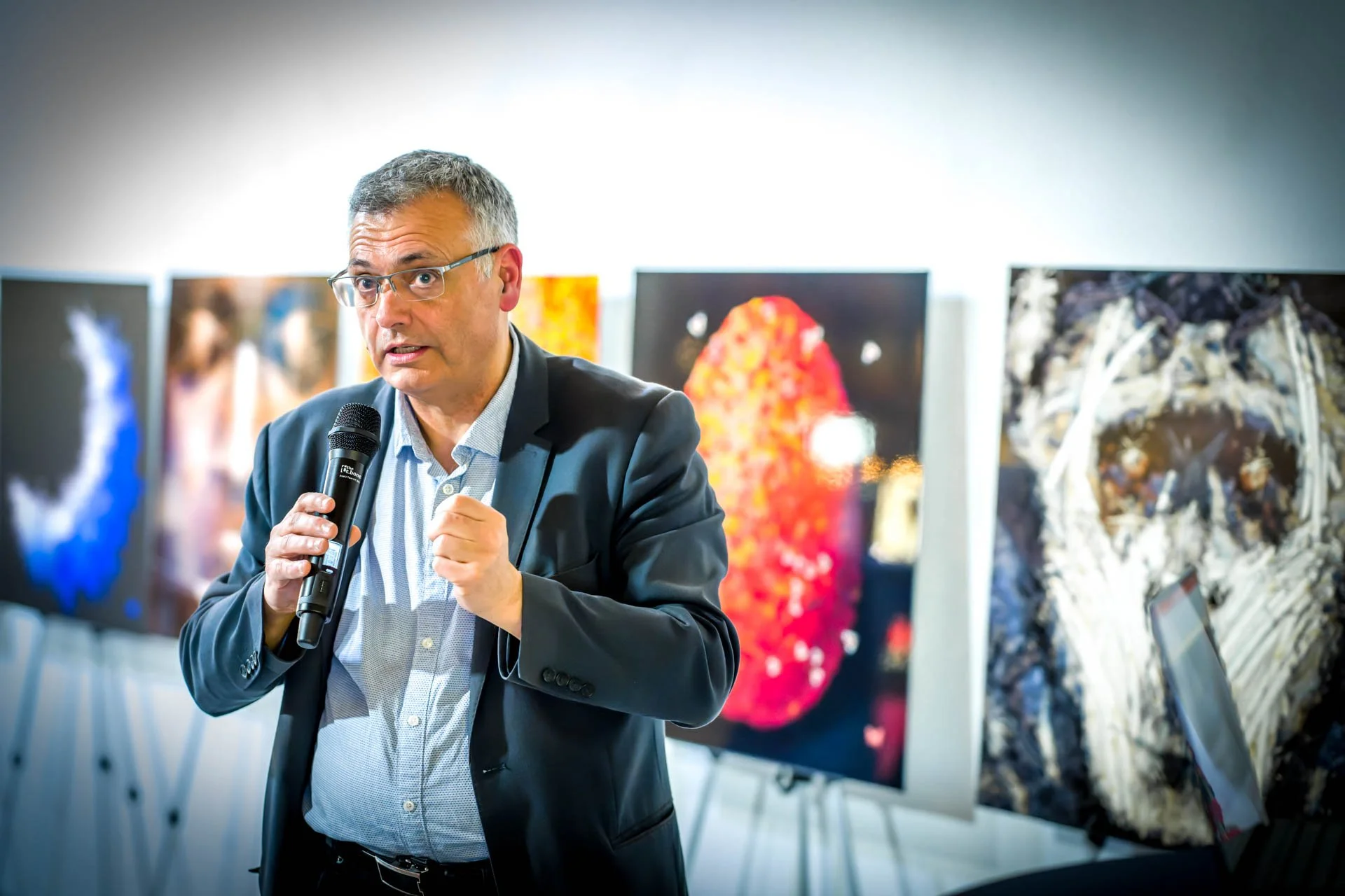 A man wearing a black suit speaks during a presentation or lecture in an art gallery, with artistic images in the background.