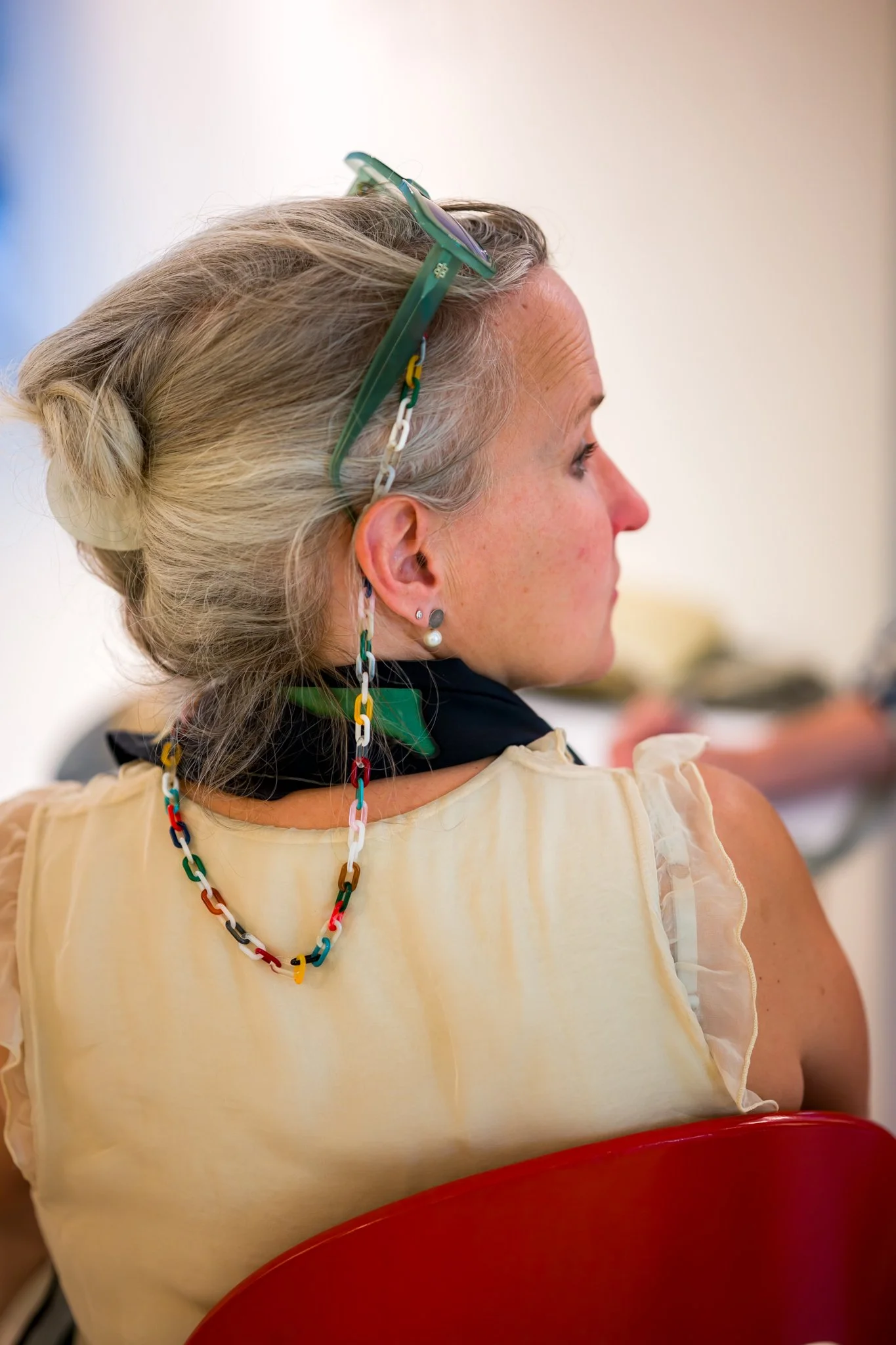 Une femme aux cheveux gris coiffés en chignon, portant des lunettes vertes sur sa tête, des boucles d'oreilles et un collier coloré en chaîne, assise sur une chaise rouge, vue de profil.