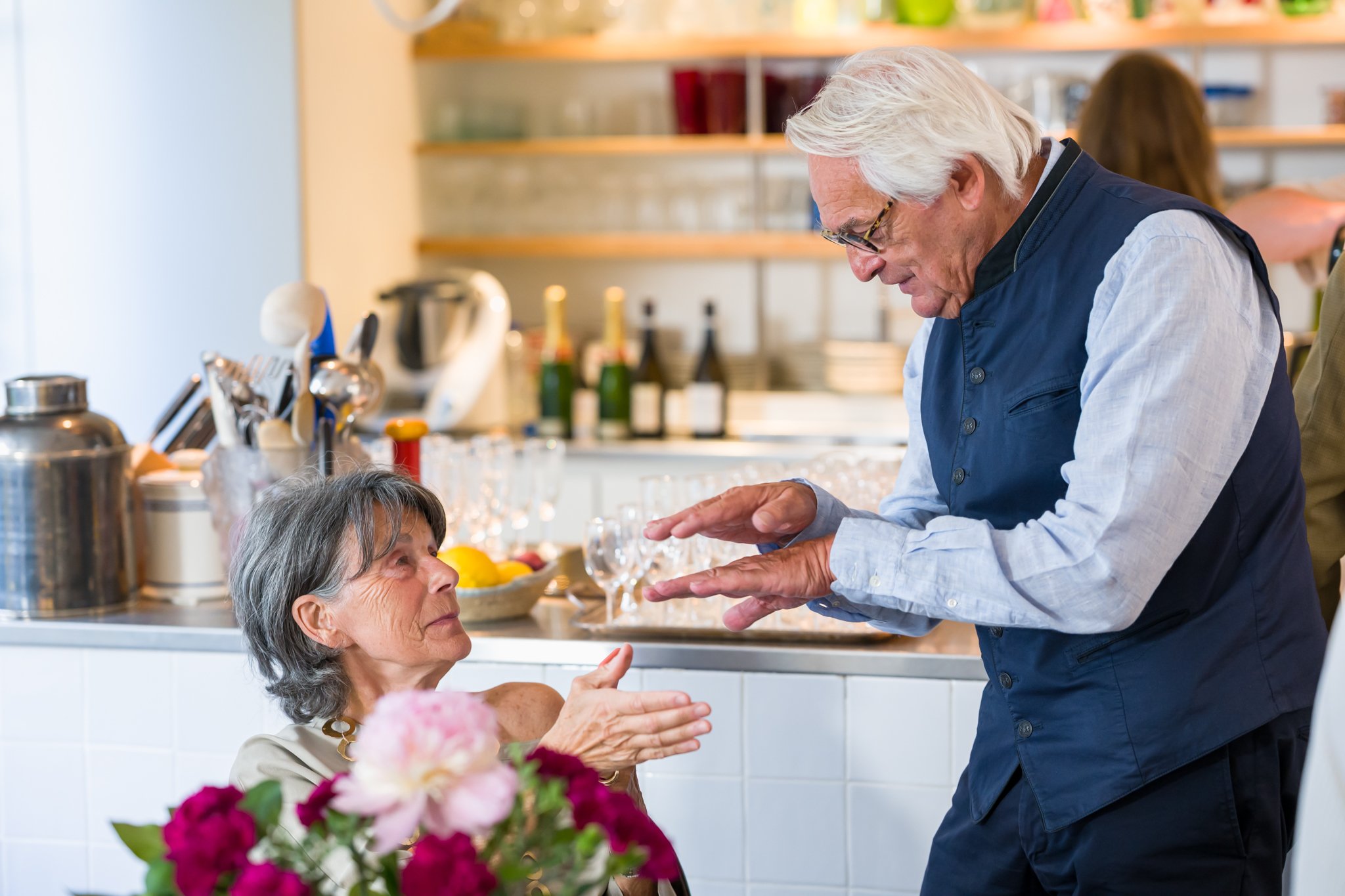 Un homme âgé avec des cheveux blancs, portant une veste bleue, semble parler ou raconter quelque chose à une femme âgée assise à une table, dans une cuisine moderne