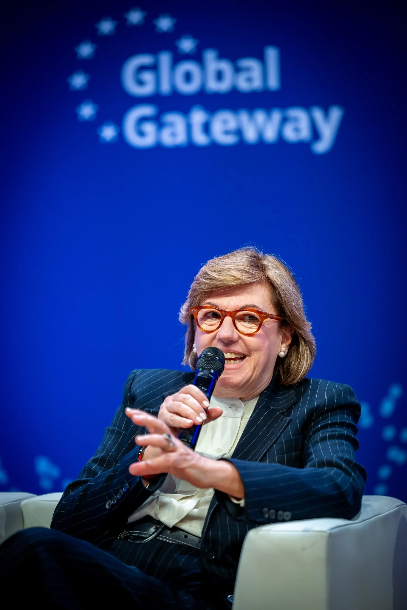 A woman smiling and speaking into a microphone, wearing glasses and a striped suit. The blue background displays the words "Global Gateway" with stars around them.