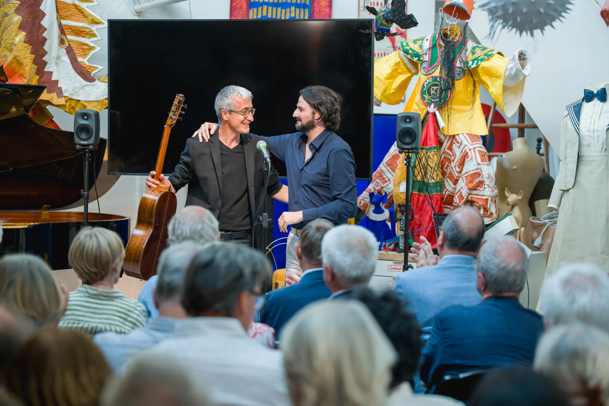 Deux hommes se saluant chaleureusement lors d'un événement, un avec une guitare. Audience attentive, décor coloré avec des sculptures, costumes et un grand écran en arrière-plan.