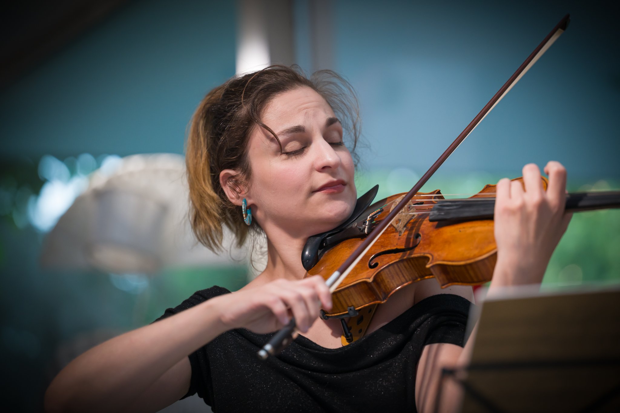 Femme jouant du violon avec yeux fermés, cheveux courts, portant des boucles d'oreilles turquoise, dans un environnement artistique.