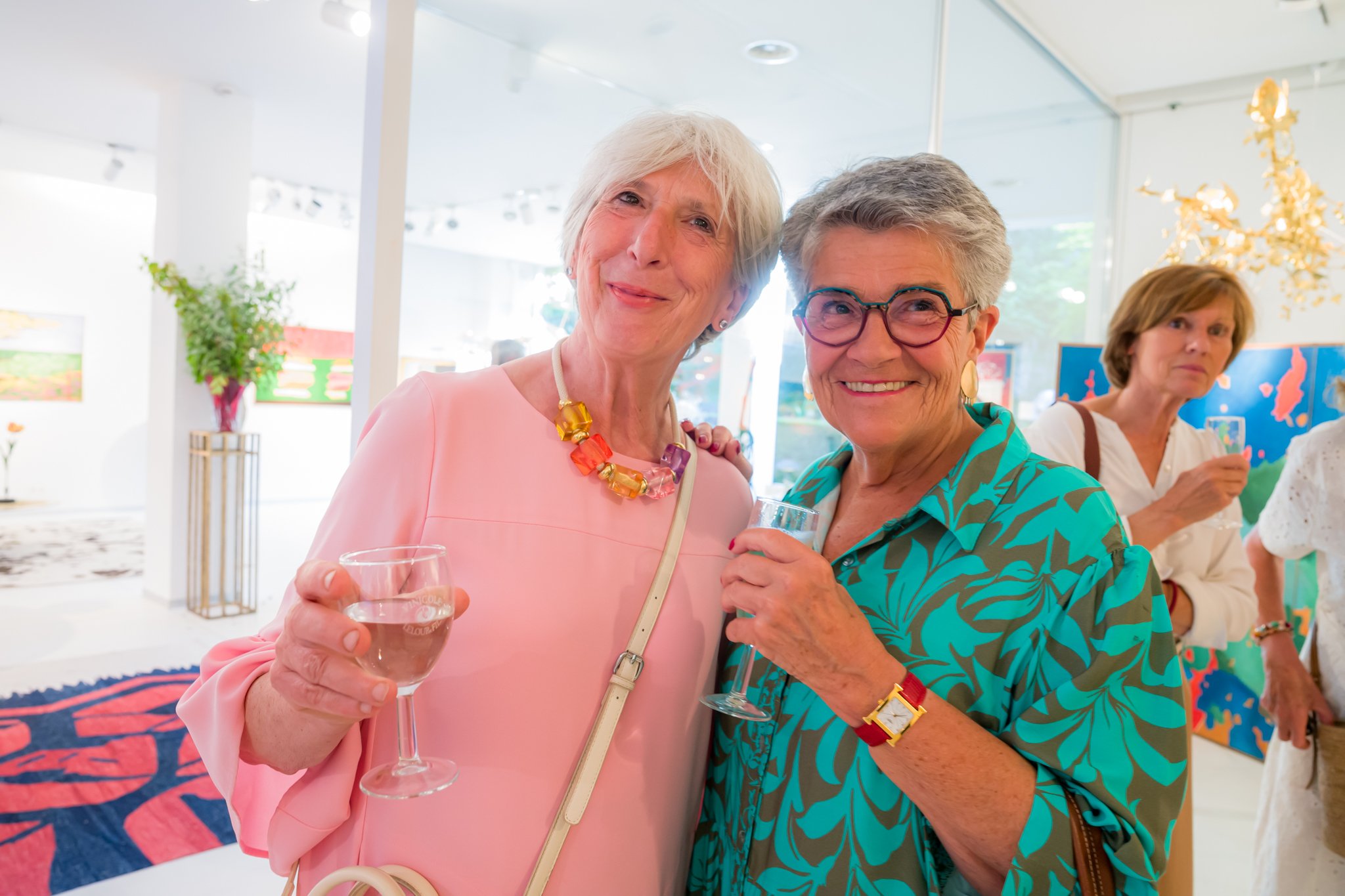 Deux femmes âgées souriantes tiennent des verres de vin lors d'une réception ou d'une fête en intérieur, avec d'autres personnes en arrière-plan.