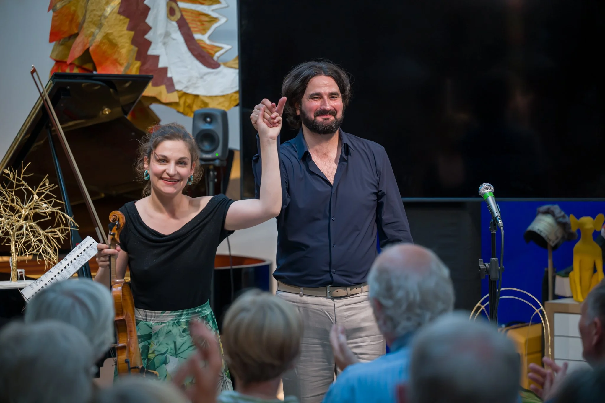 Deux musiciens saluant un public lors d'un spectacle, une femme avec un violon et un homme avec une chemise bleue, dans un lieu décoré avec des œuvres d'art colorées.