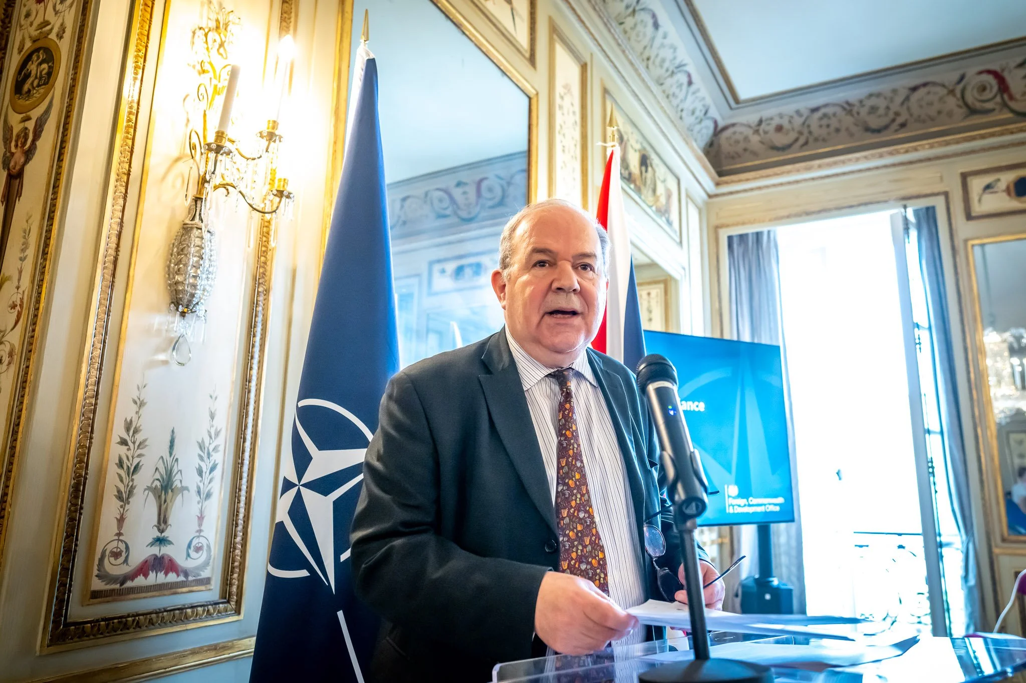A man in a suit delivers a speech at a microphone, with a NATO flag in the background in a decorated room.