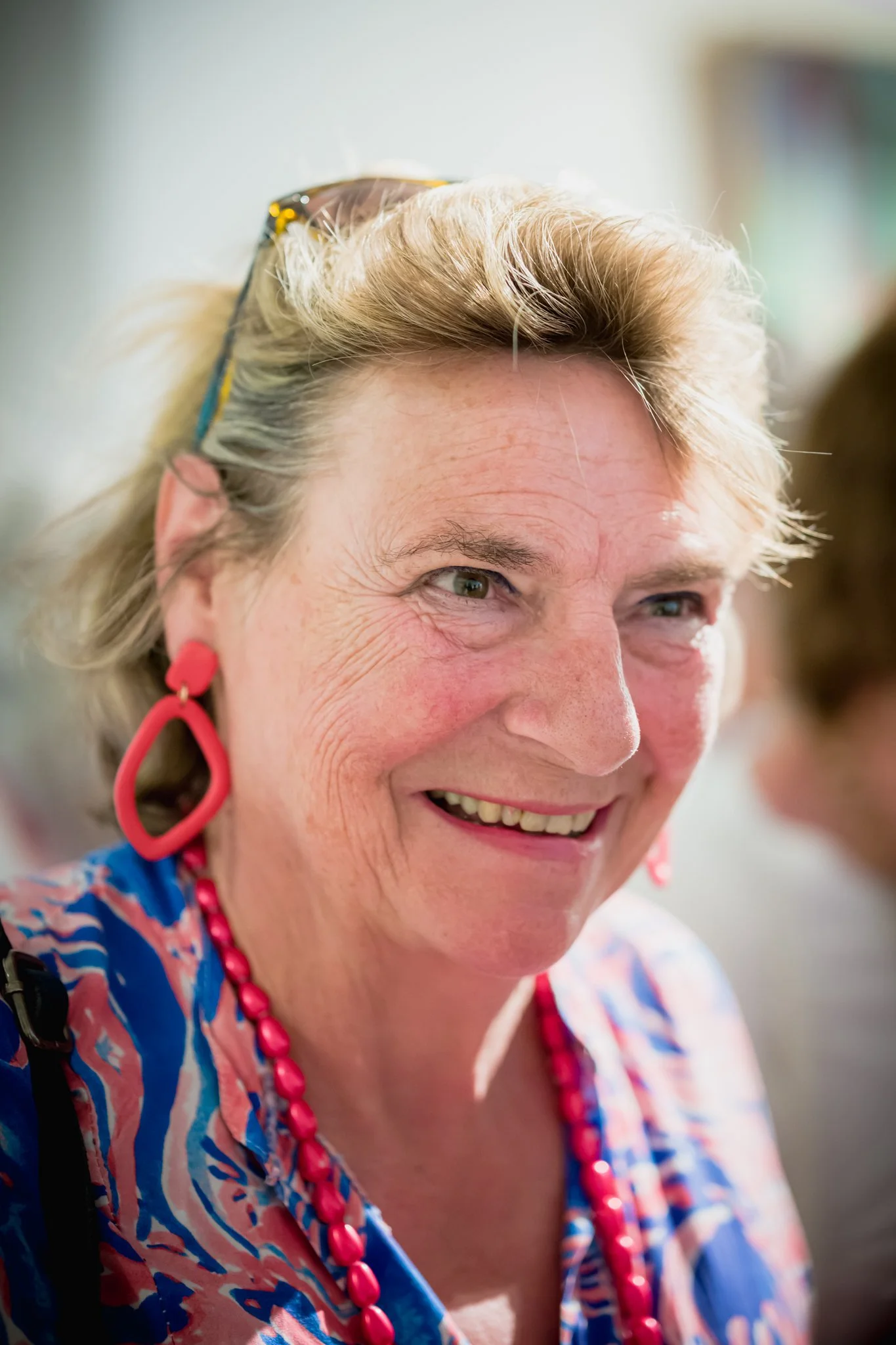 Une femme âgée souriante portant de grosses boucles d'oreilles rouges et un collier de perles roses, habillée en couleurs vives, dans un environnement lumineux.