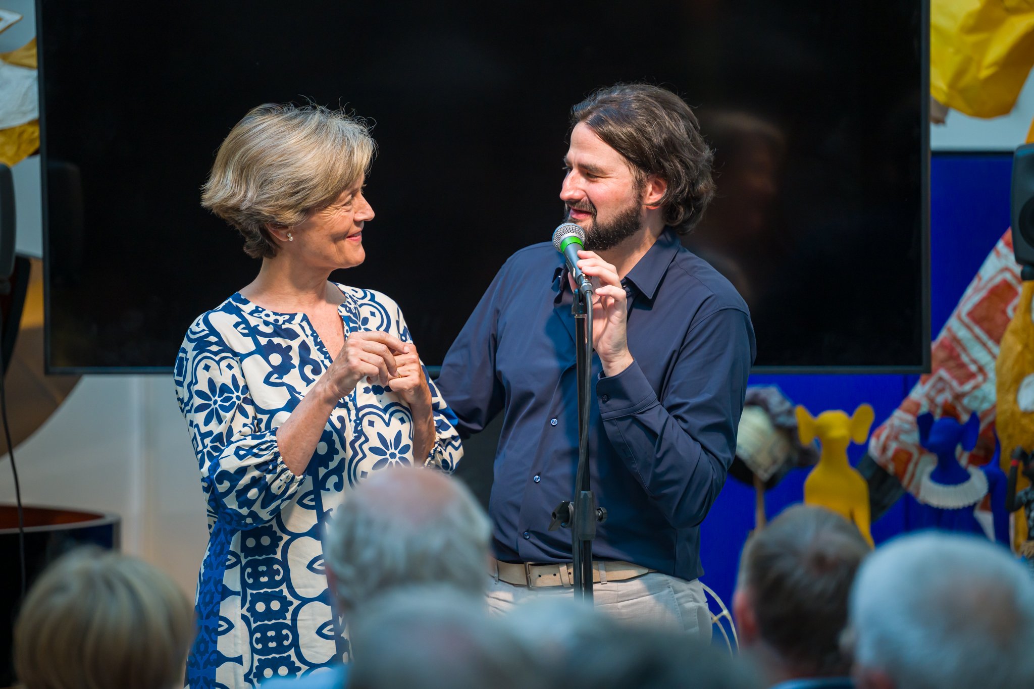 Une femme âgée en robe blanche et bleue avec un motif floral, souriante, et un homme avec une barbe, en chemise bleue, parlant dans un micro devant un public.