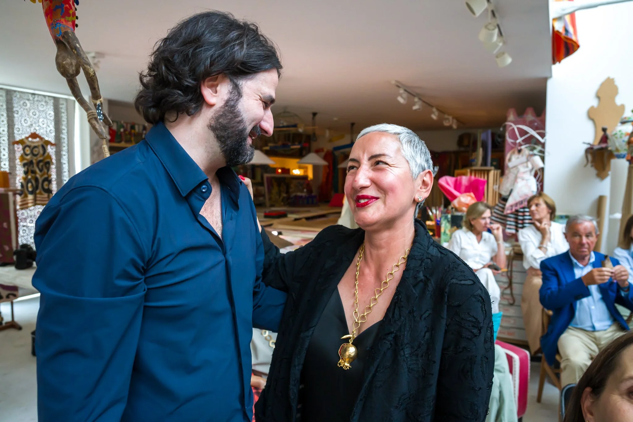 Un homme et une femme sourient et semblent échanger des plaisanteries lors d'une réunion ou d'un événement social à l'intérieur. Autres personnes en arrière-plan, quelques objets colorés et décorations artisanales.