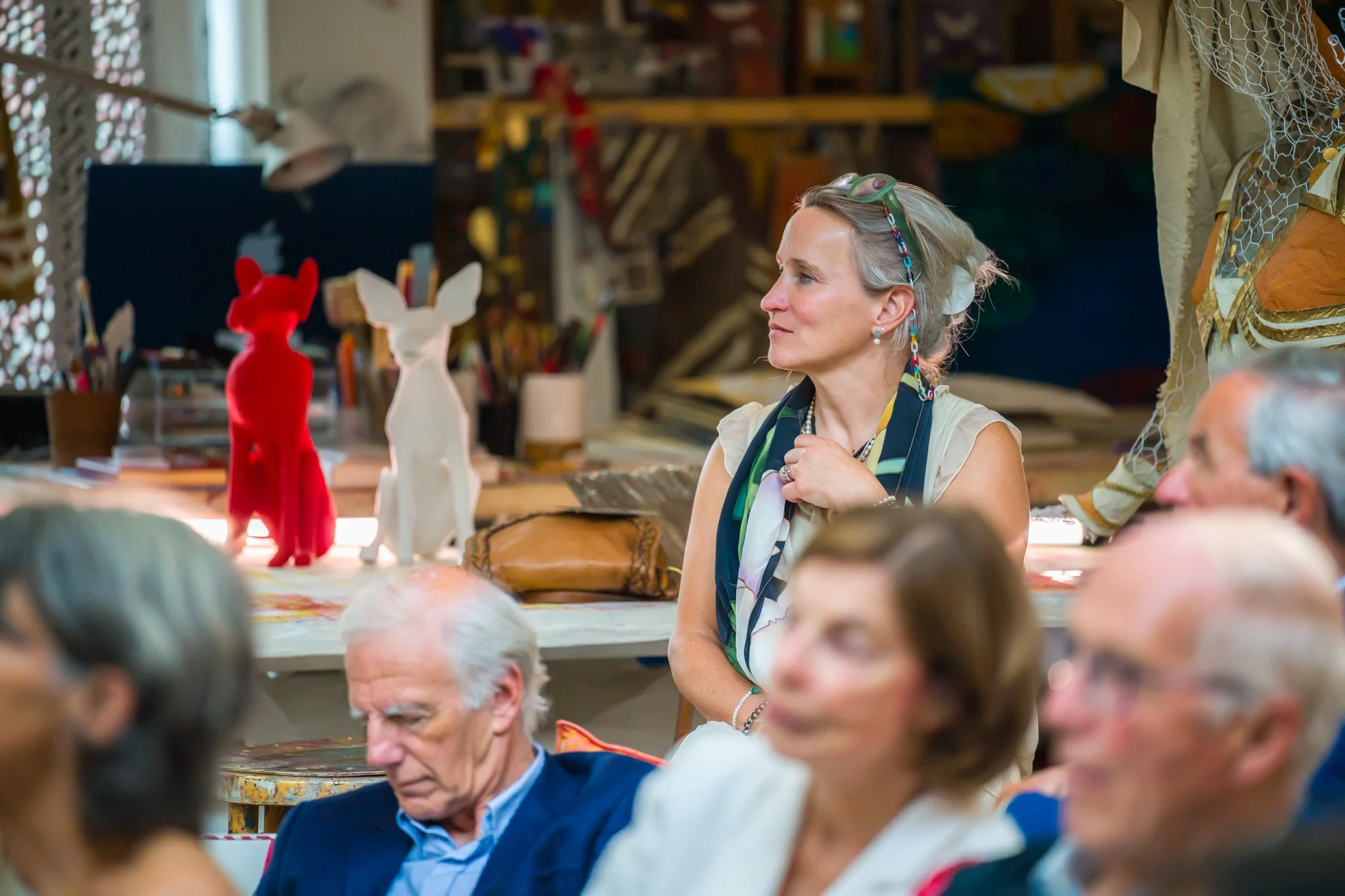 Une femme assise observe attentivement une présentation ou une discussion dans une salle, entourée de plusieurs personnes. Derrière elle, deux figurines en forme de lapin ou de renard, une rouge et une blanche, sont visibles sur une table.