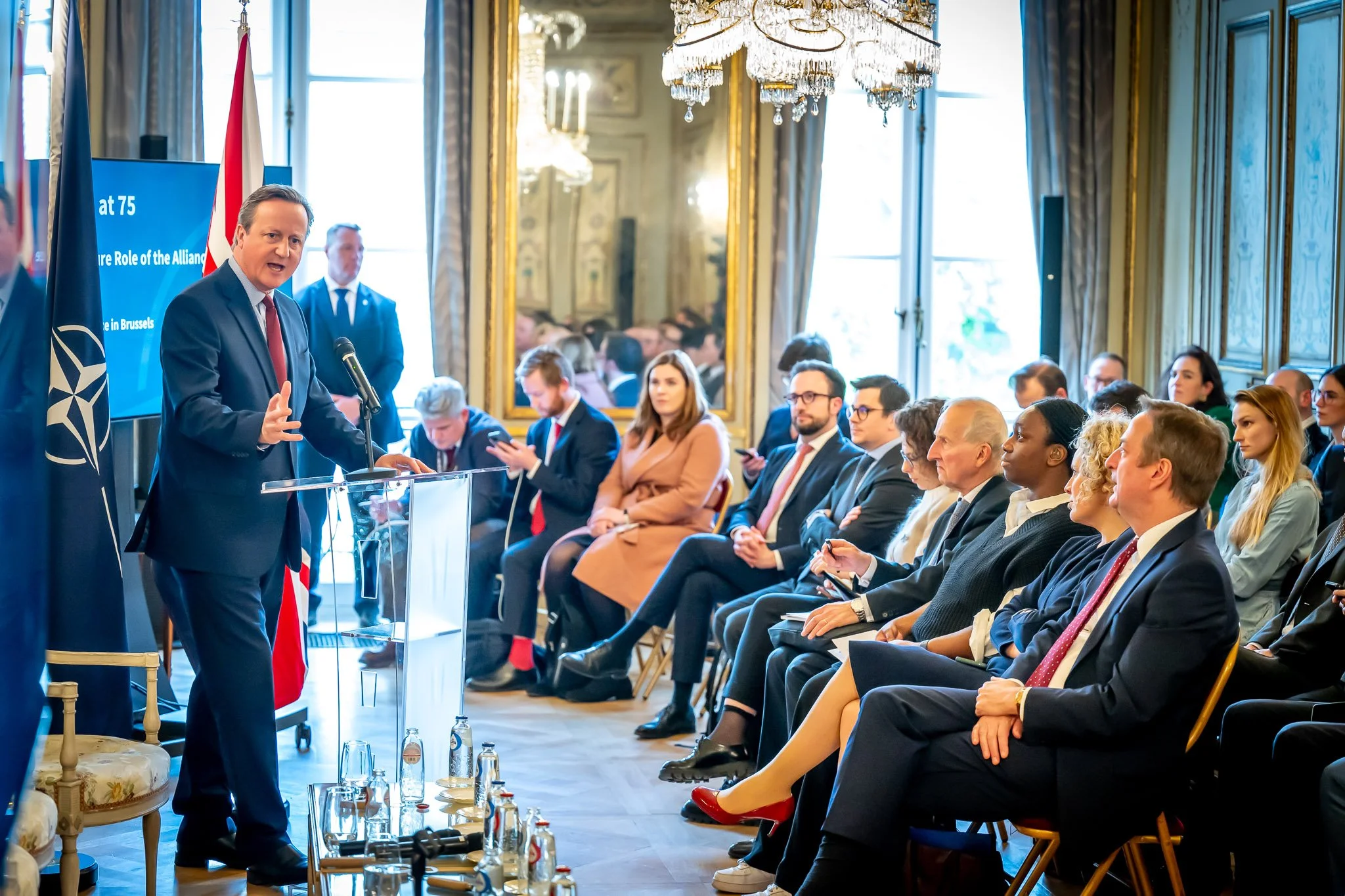 A man in a suit speaks to an audience at a conference, with a NATO flag and screen in the background. The participants are attentive and seated in an elegant room with a chandelier.