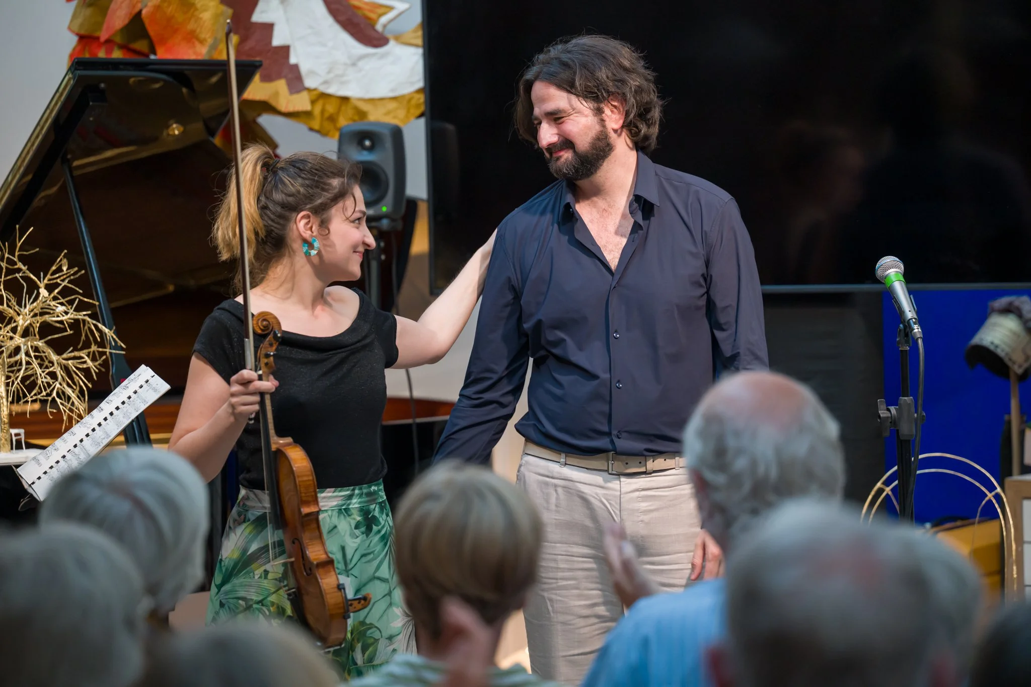 Une femme et un homme se tiennent face à face sur scène, la femme tient un violon et serre l'épaule de l'homme, dans une ambiance de performance ou de concert, avec un public d'audience devant eux.