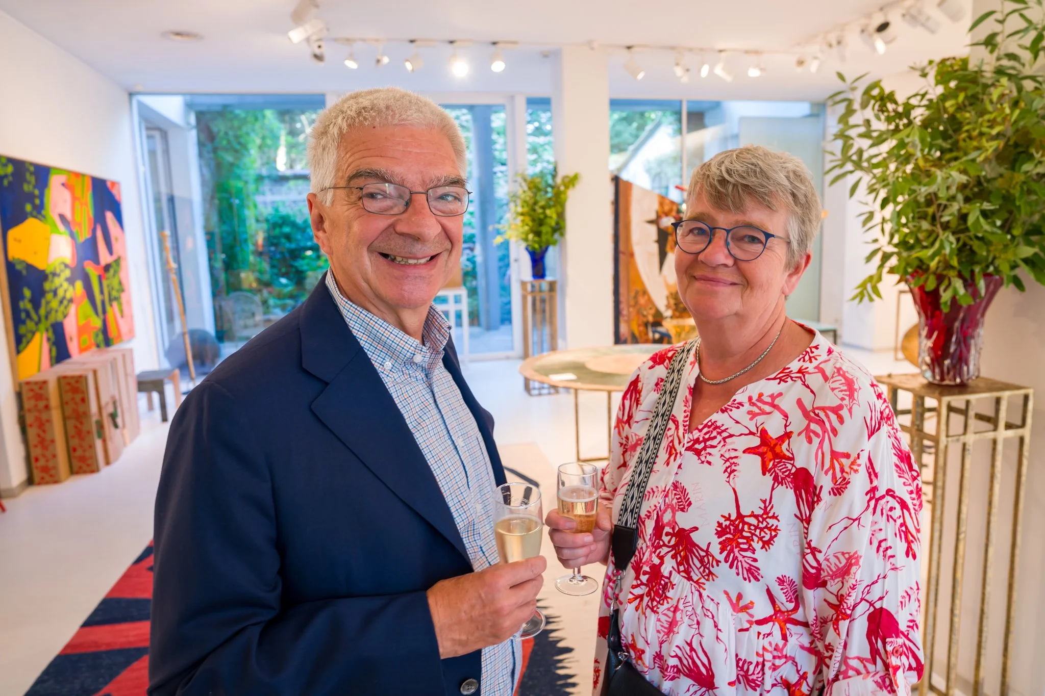 Deux personnes souriantes, un homme en costume bleu et une femme à lunettes avec une chemise aux motifs rouges et blancs, tenant des verres de champagne, lors d'une réception ou d'une fête dans un espace d'art contemporain avec des peintures et une g