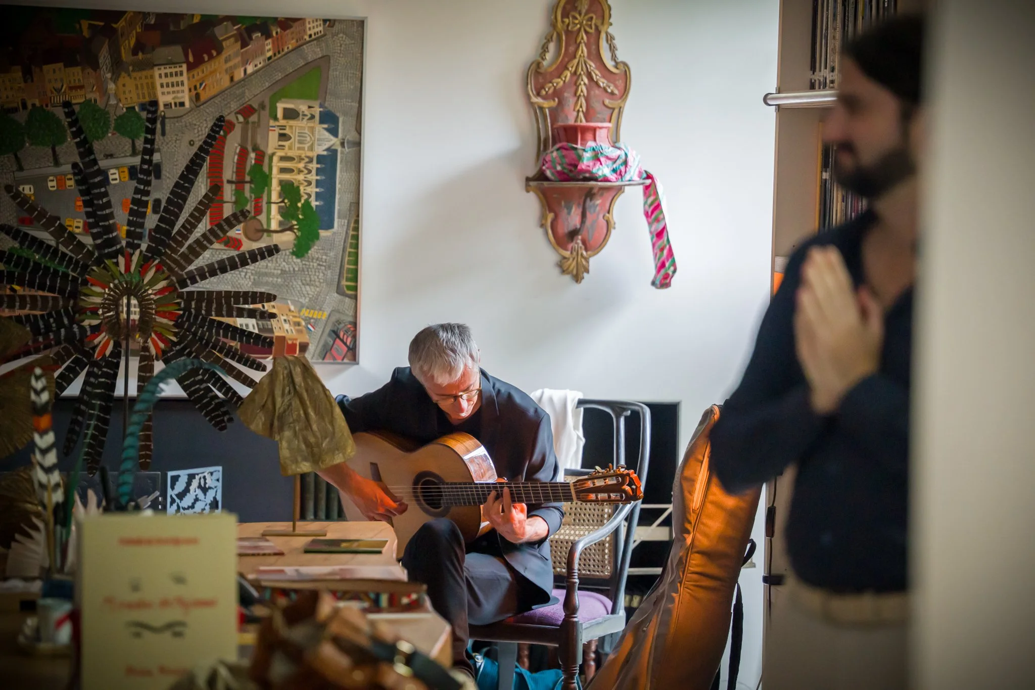 Un homme joue de la guitare dans une pièce décorée avec un tableau coloré et une sculpture murale, tandis qu'un autre homme se tient près d'une porte, la main sur la poitrine.