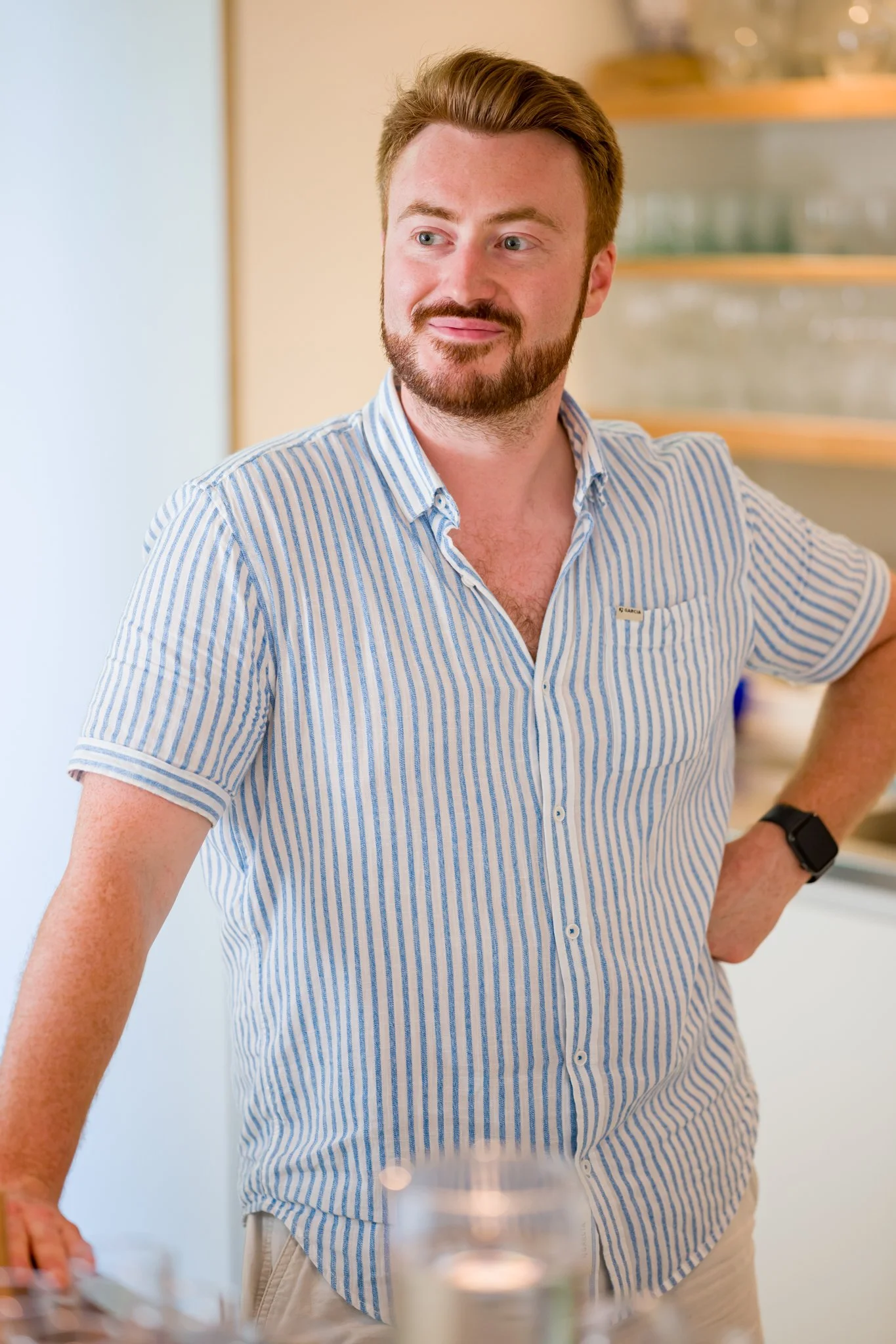 Homme avec barbe, portant une chemise à rayures bleues et blanches, souriant, posé dans une cuisine