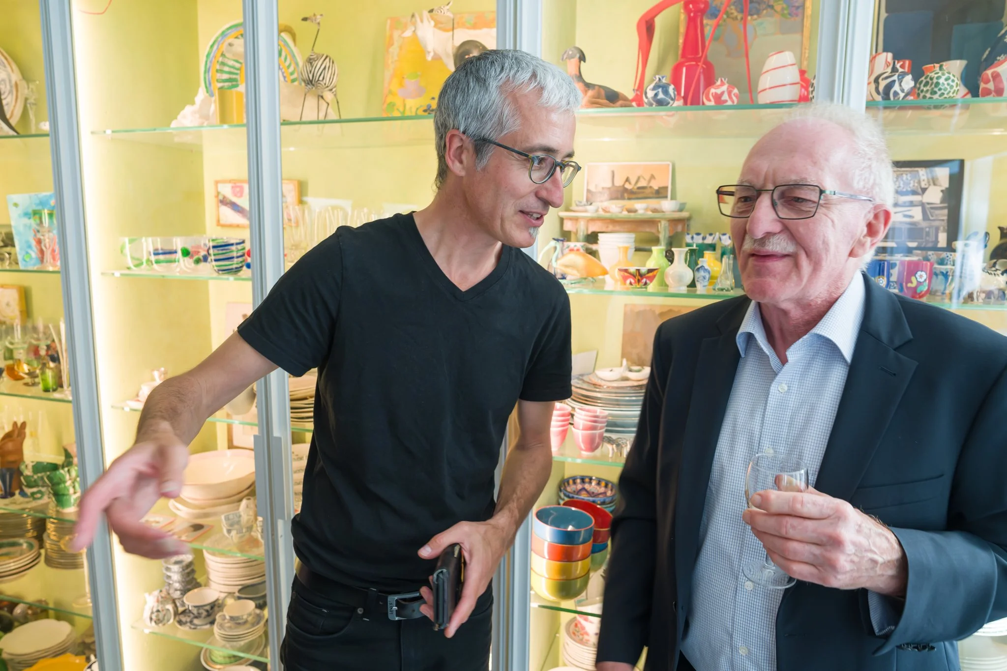 Deux hommes discutent dans une boutique d'articles en céramique, entourés d'étagères remplies de vaisselle colorée.