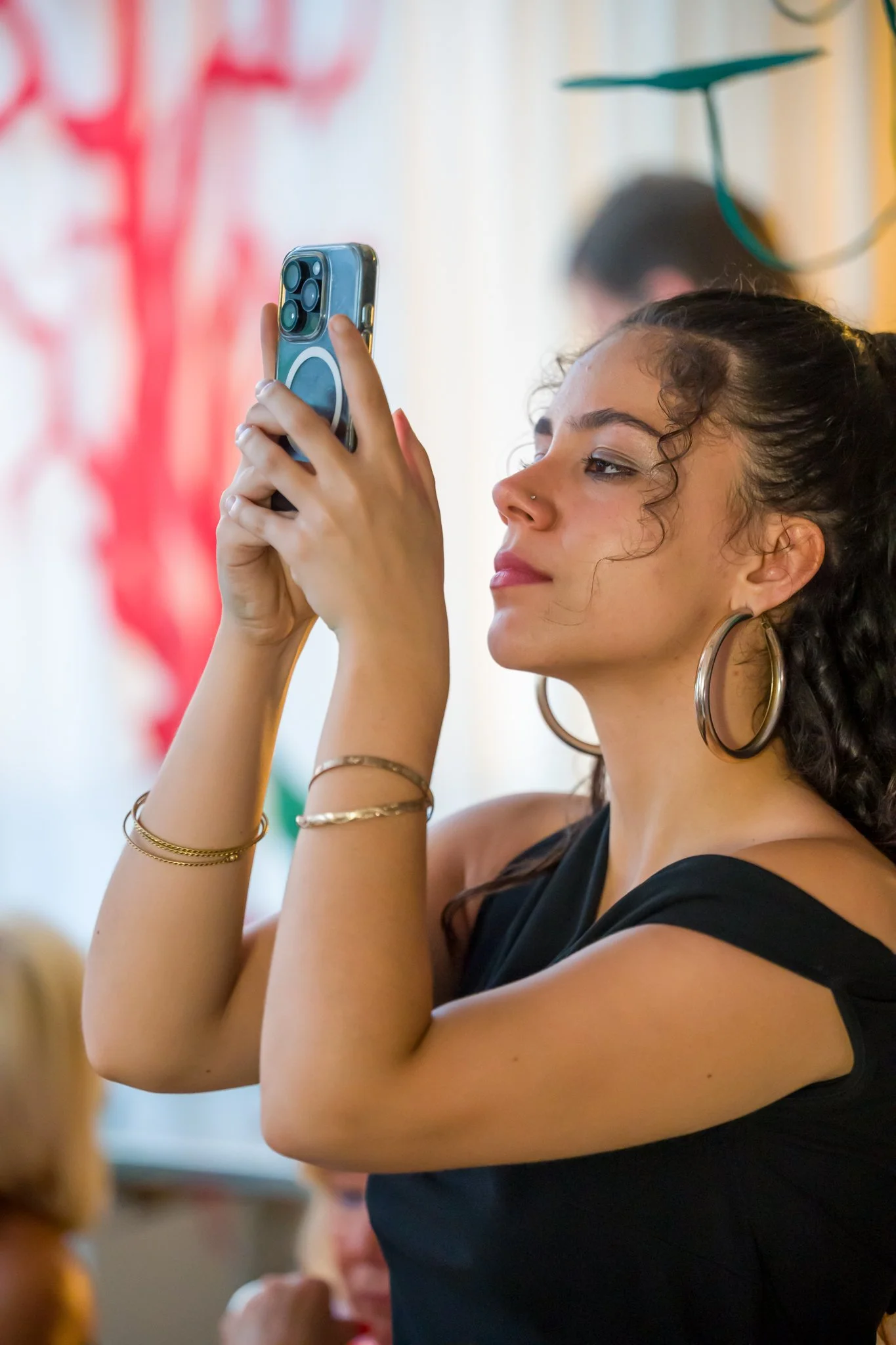 Jeune femme regardant son téléphone portable, portant de grands boucles d'oreilles en métal, bracelets, et une robe noire, dans un lieu décoré avec des couleurs vives.