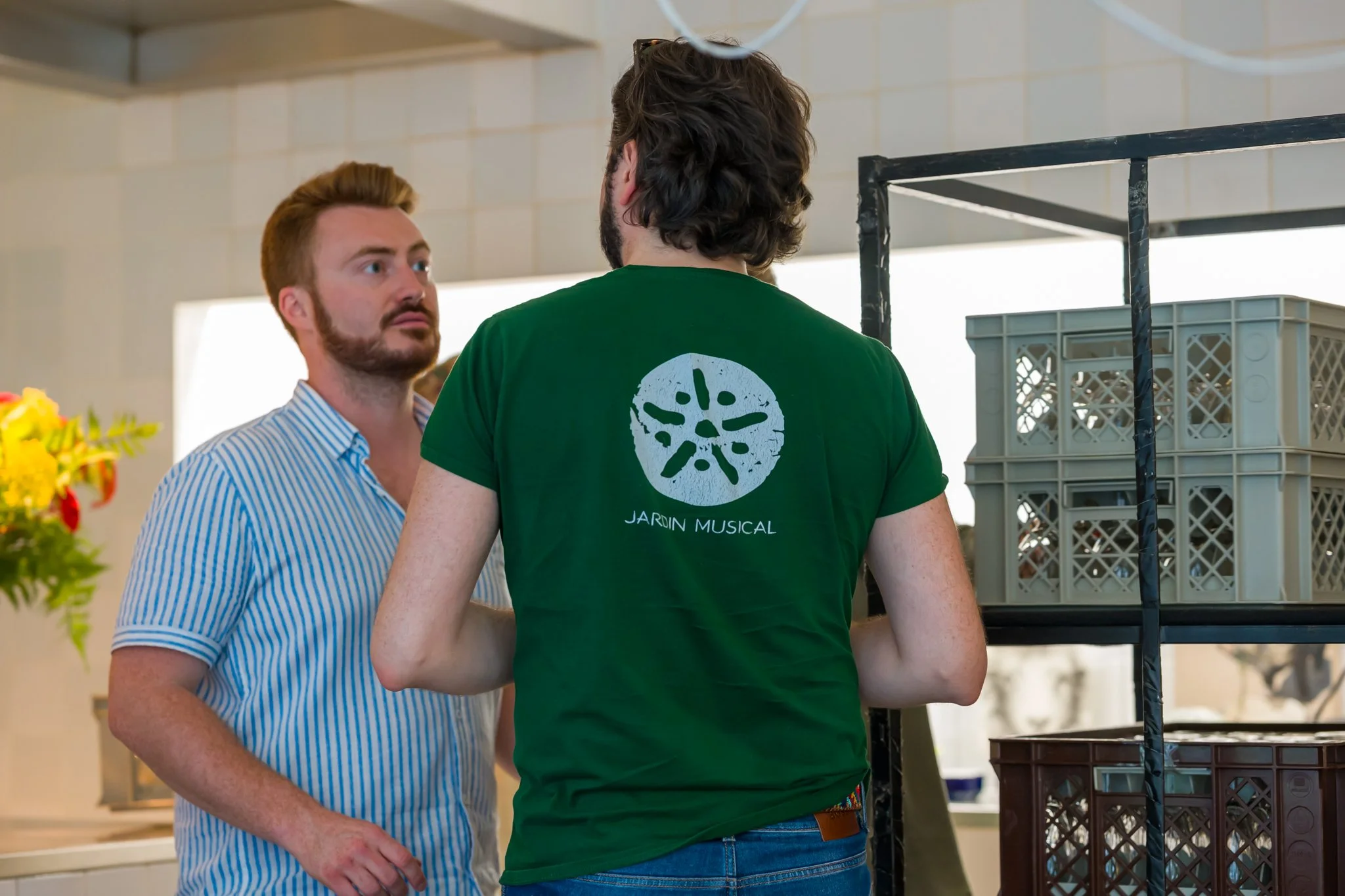 Deux hommes discutent dans une cuisine, l'un porte une chemise à rayures et l'autre un t-shirt vert avec l'inscription "Jardin Musical" et un symbole graphique.