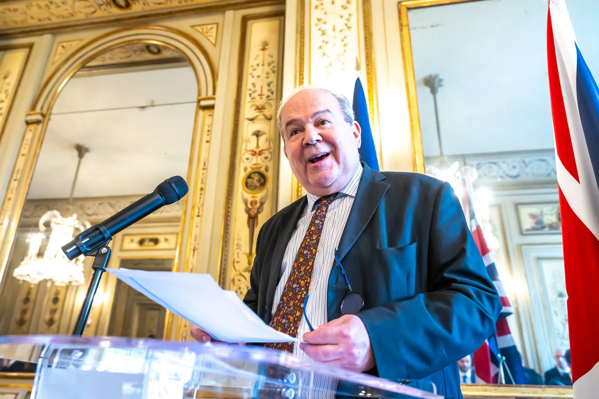 Smiling man holding papers behind podium with microphone, flags in background.