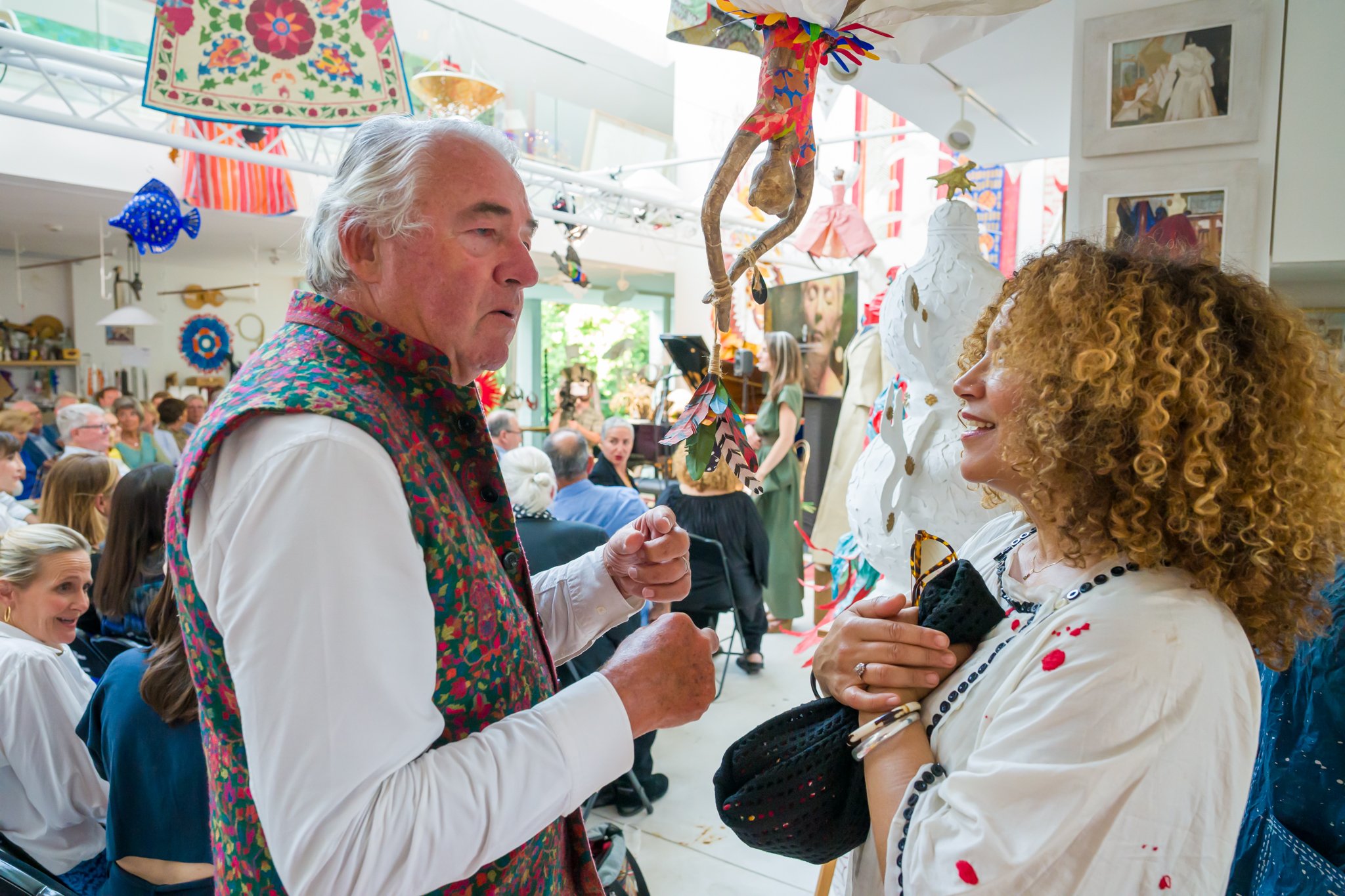 Deux personnes discutent lors d'une fête ou d'un événement artistique, avec des décorations colorées et une foule en arrière-plan.