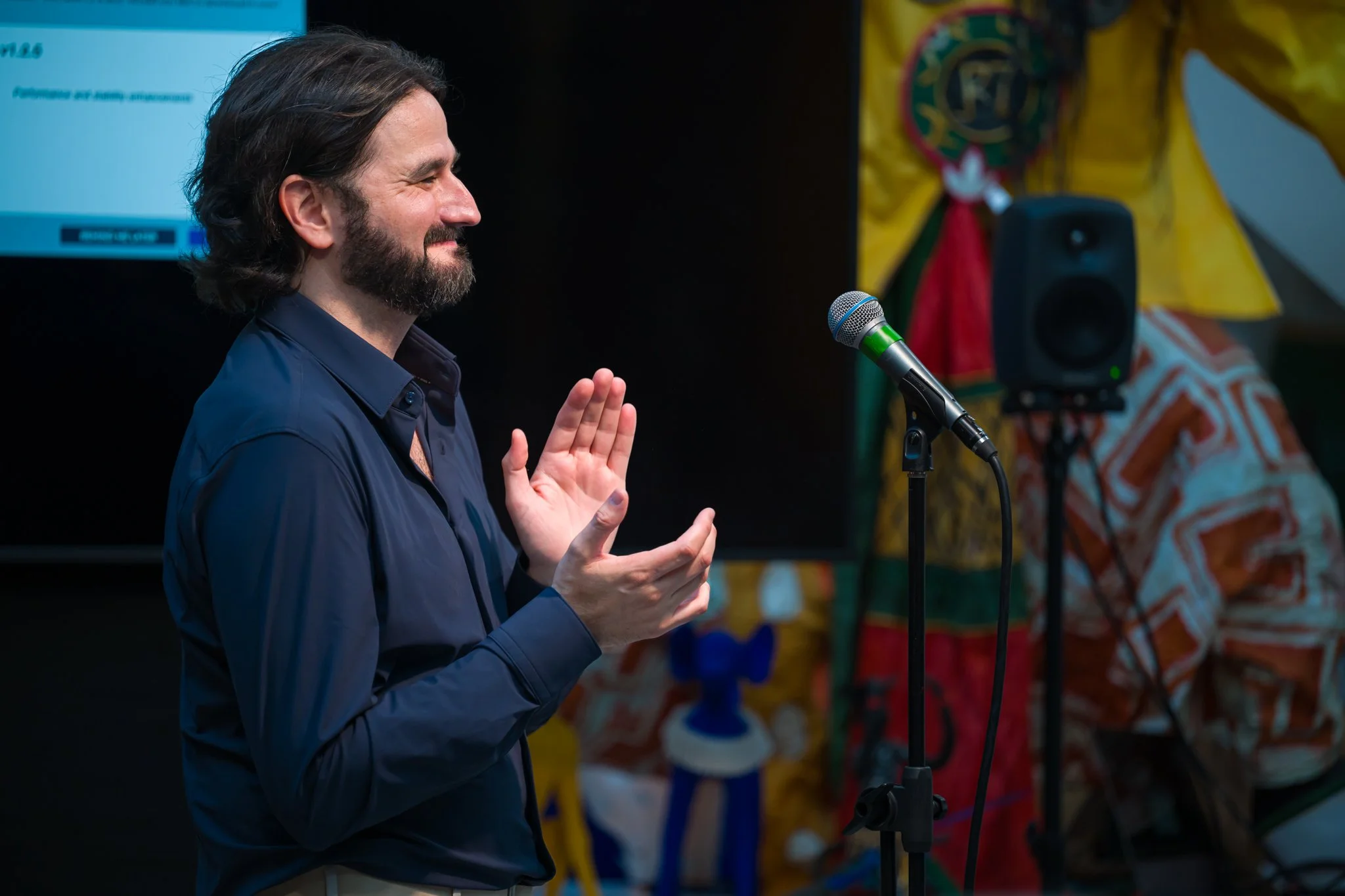 Homme avec barbe et cheveux longs, souriant et levant une main, parlant devant un micro lors d'une présentation.