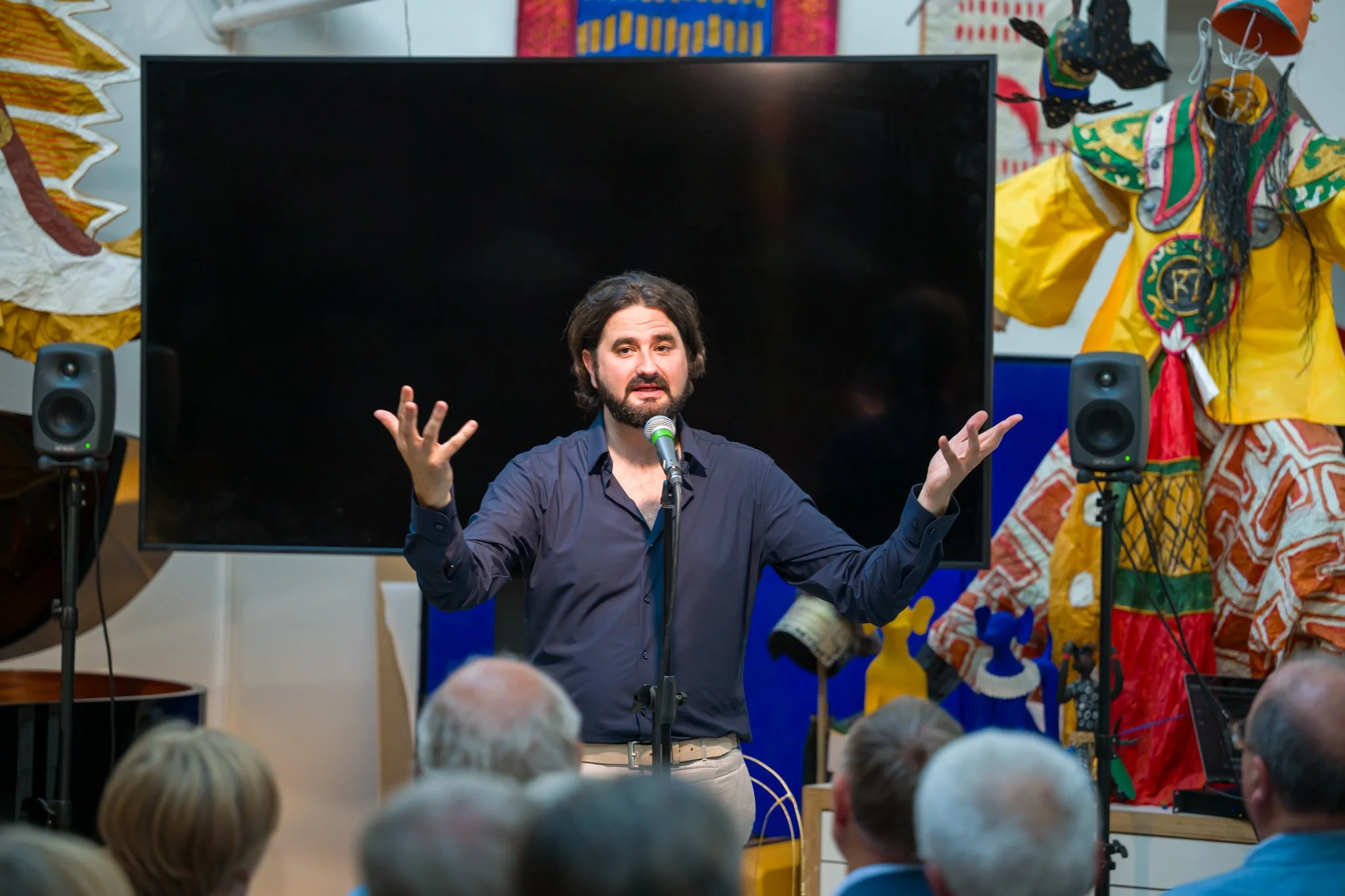 Un homme parle devant un micro lors d'une présentation ou d'un spectacle, avec un écran noir derrière lui et des décorations colorées autour.