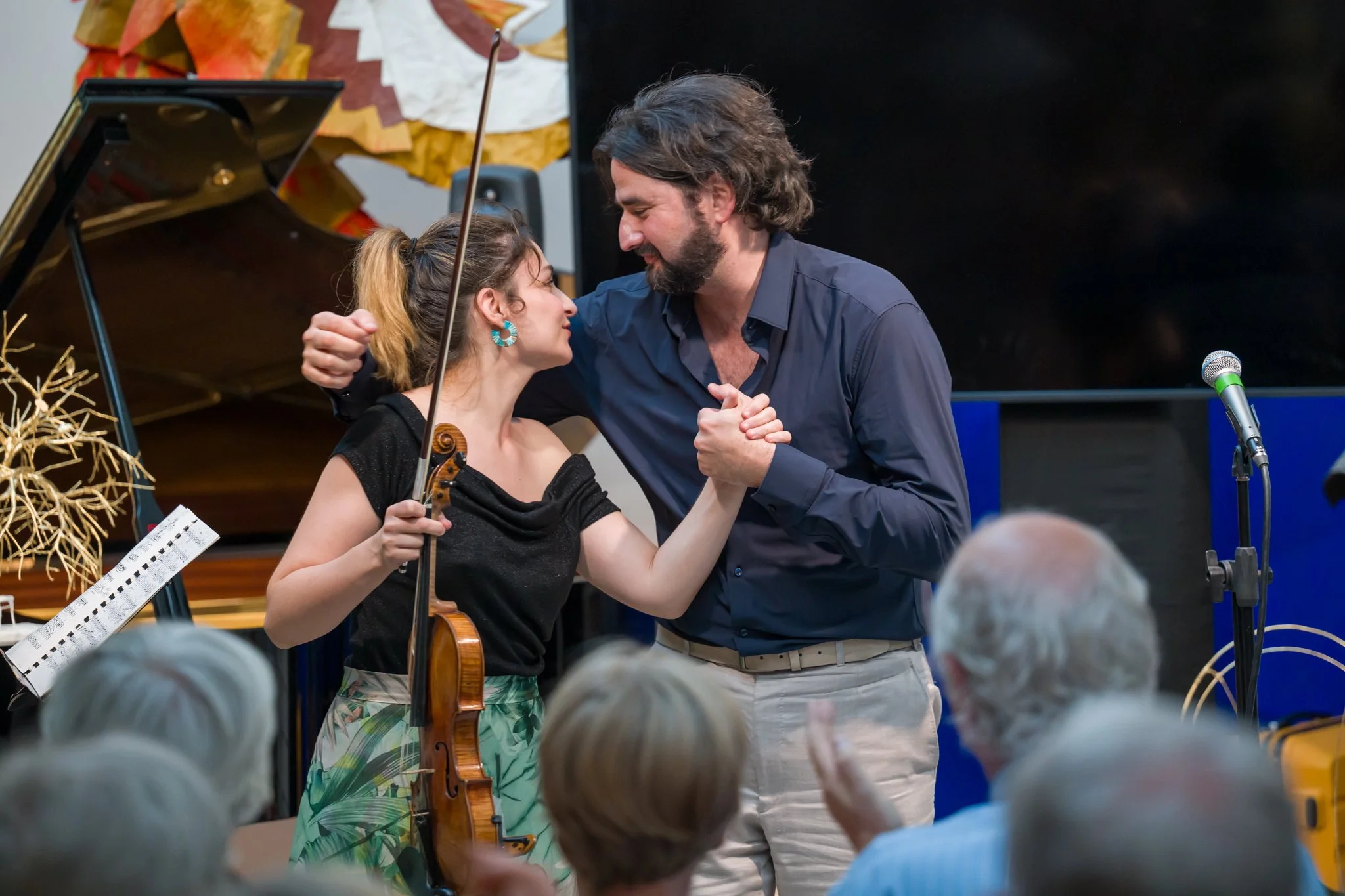 Une jeune femme et un homme se tiennent la main lors d'une performance musicale, entourés d'un public. La jeune femme porte un violon et une robe à motifs verts, et semble recevoir une reconnaissance ou un hommage.