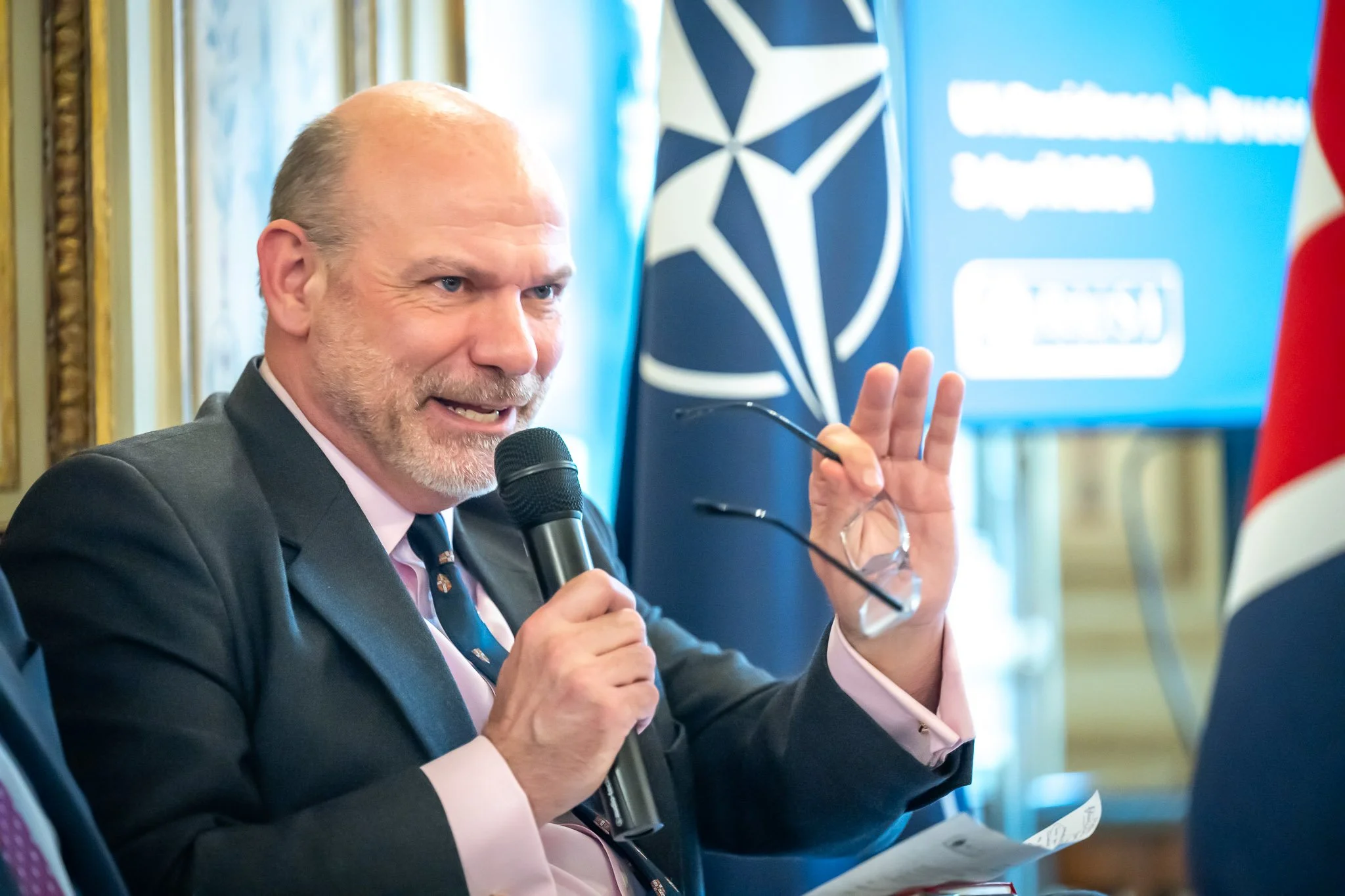 Man holding a microphone and speaking at an event with a NATO flag and logo in the background.